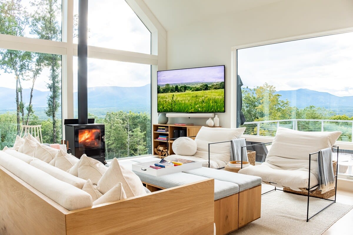 Cozy living room with a light wood sofa and armchairs, a wall-mounted TV, and a wood-burning stove, all set against a backdrop of large windows showcasing mountain and forest views.
