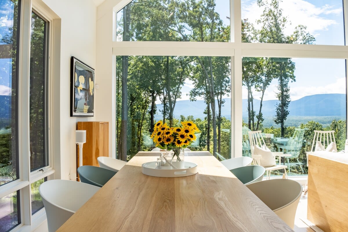 Bright dining area with a large light wood table and modern white chairs, set against a backdrop of expansive floor-to-ceiling windows offering stunning forest and mountain views.