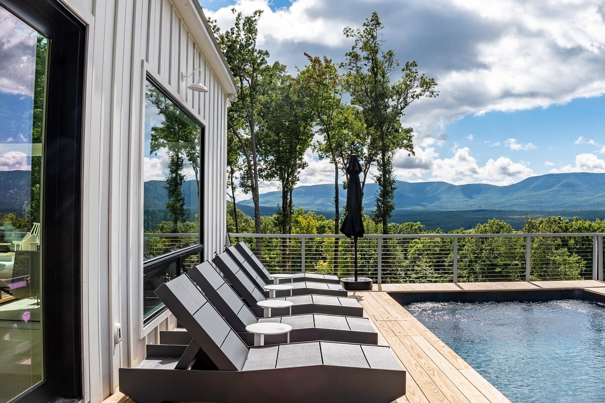 A modern swimming pool area on a wooden deck, lined with dark grey lounge chairs and small side tables. The pool overlooks a breathtaking panorama of rolling mountains and lush trees under a partly cloudy sky.