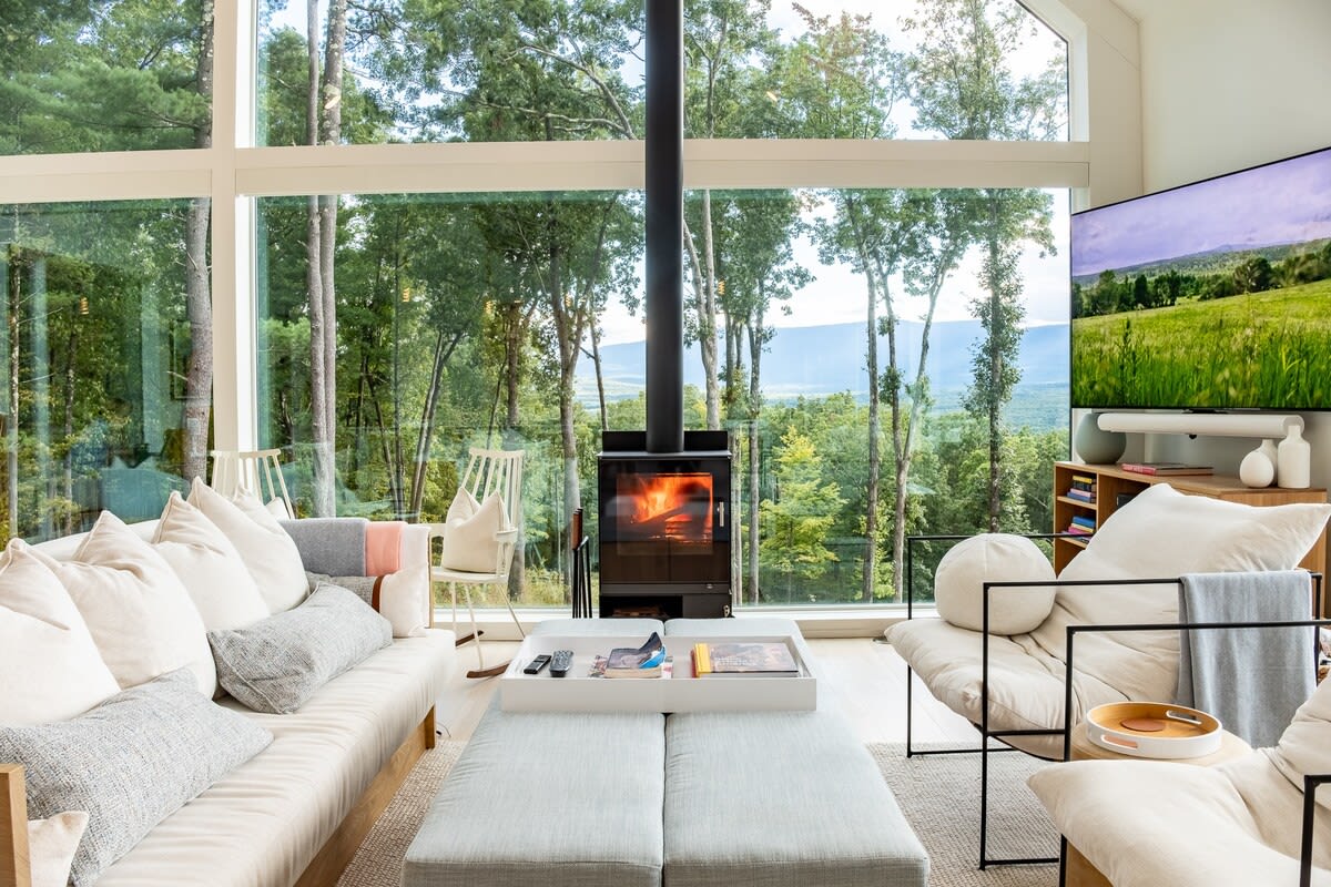 Bright living room with floor-to-ceiling windows showcasing expansive forest and mountain views, furnished with a light-colored sofa, a low ottoman coffee table, and a modern wood-burning stove.