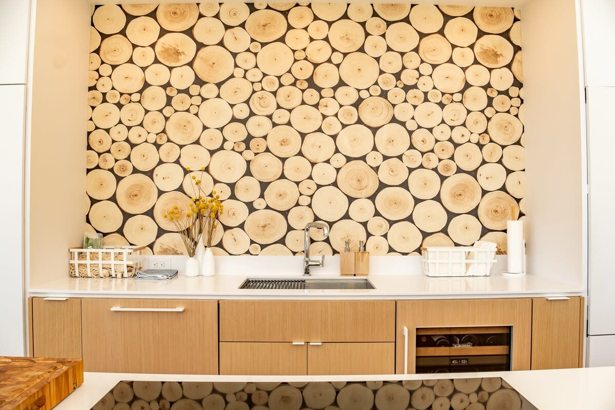 Detailed view of the modern kitchen's sink area, showcasing the wood slice accent wall, white countertops, and light wood cabinetry, with a built-in wine cooler visible.