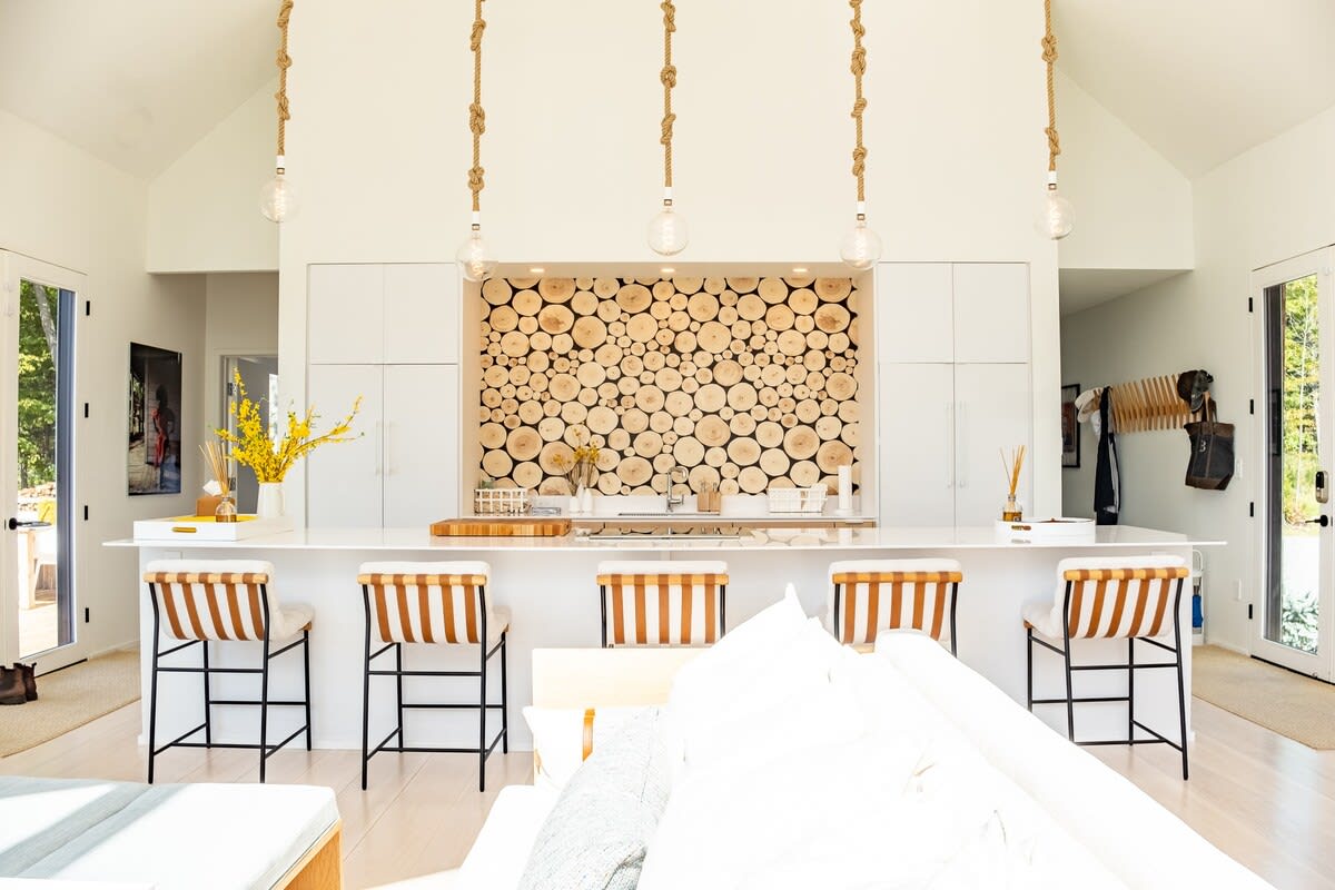 Open-concept kitchen with a large island and breakfast bar, featuring light wood cabinetry, white countertops, and a striking wood slice accent wall, illuminated by unique rope pendant lights.