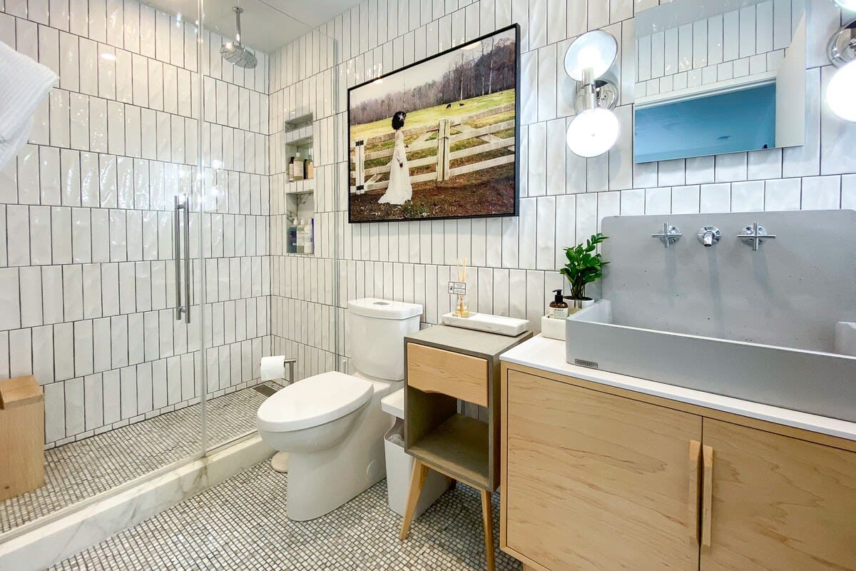 A modern bathroom with white vertical subway tiles in the shower and a patterned floor, featuring a toilet, a unique concrete-style trough sink with wall-mounted faucets, and a large framed artwork above the toilet.