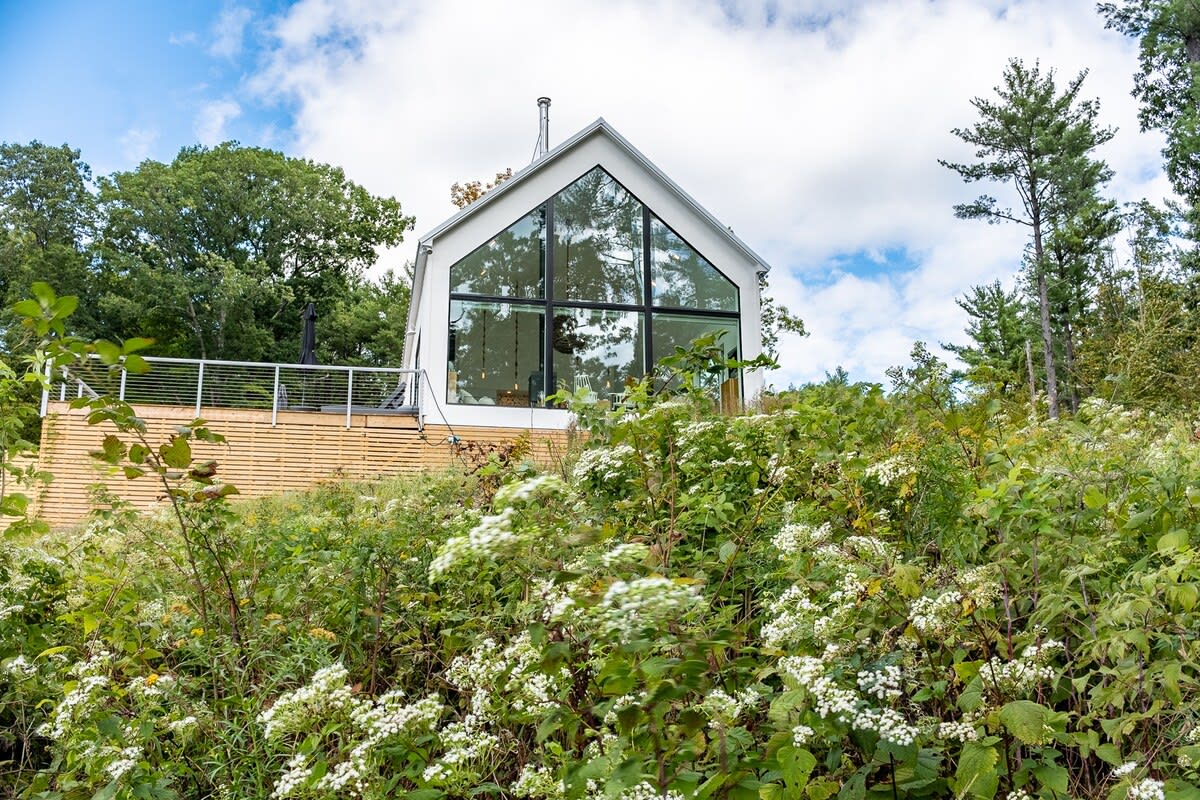 Modern white house with a gabled roof and a full-height glass facade, set amidst lush green foliage and white flowering plants, with a wooden deck visible on the left.