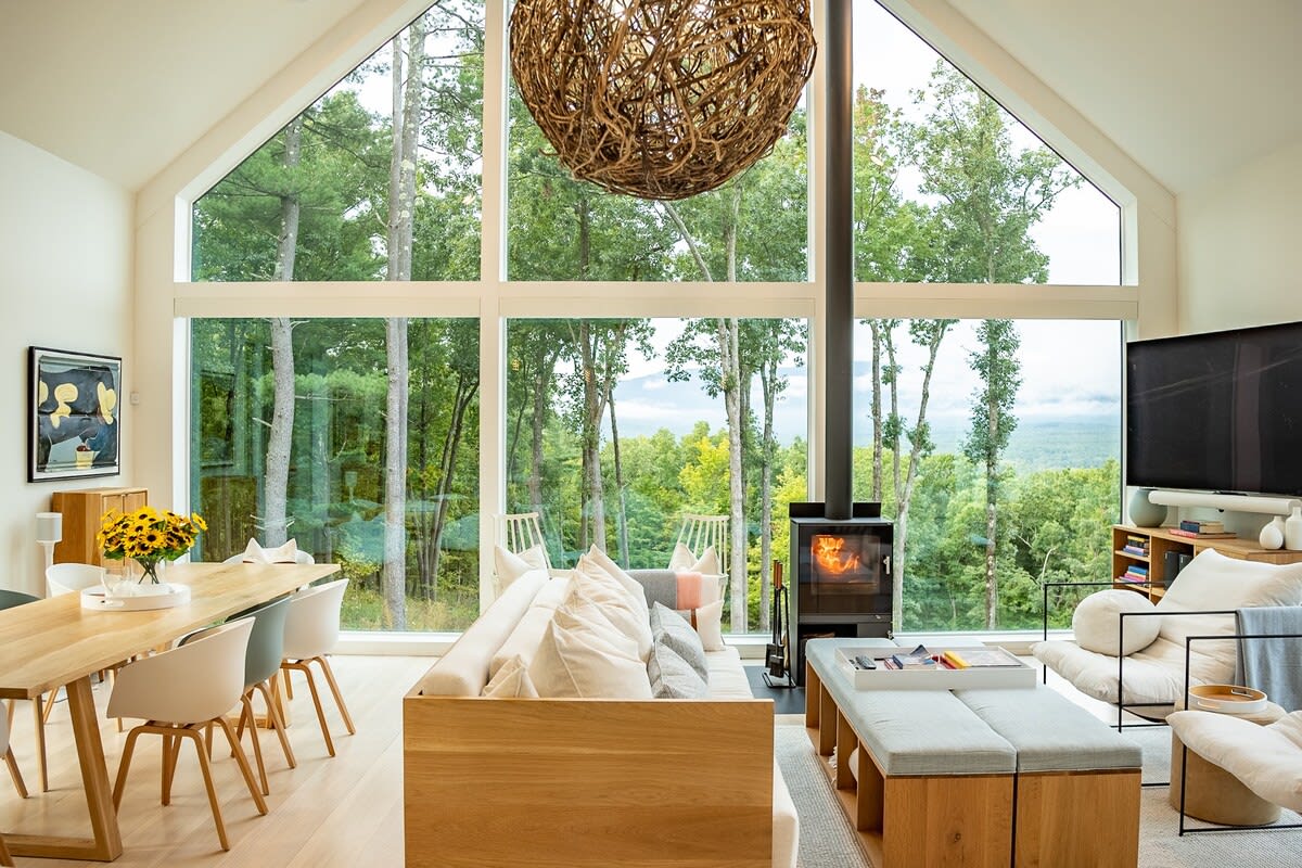 Modern open-concept living and dining area with floor-to-ceiling windows offering panoramic forest and mountain views, featuring a large wooden dining table, a contemporary sofa, and a central wood-burning stove.