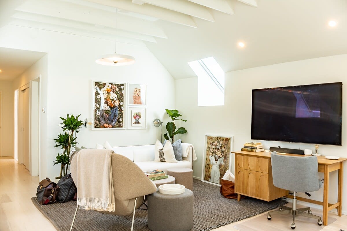 Cozy secondary living space featuring a white sofa, an armchair, a desk with an office chair, and a large wall-mounted TV, with framed floral artwork and plants adding a touch of nature.