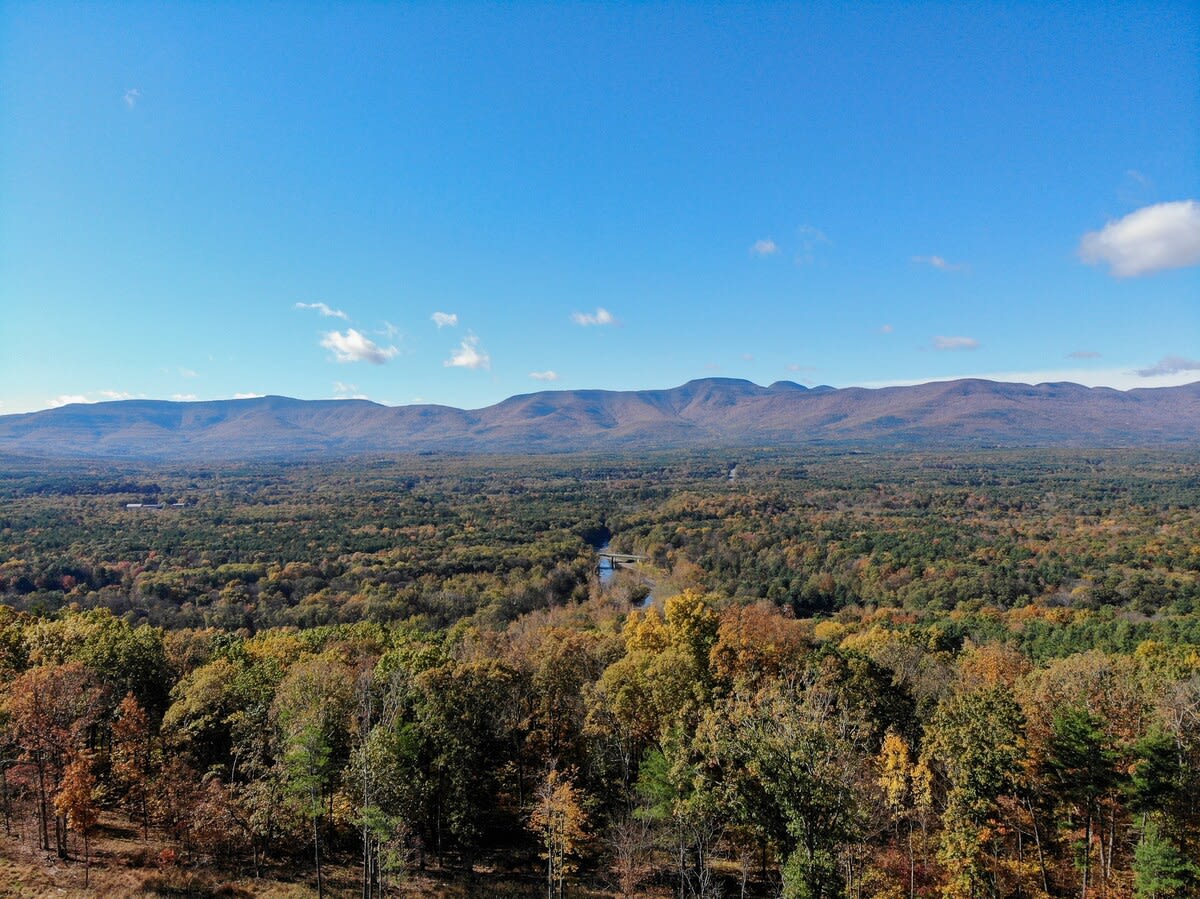 Expansive aerial view of a vast autumn landscape with a river winding through a valley of colorful trees, framed by distant rolling mountains under a clear blue sky.