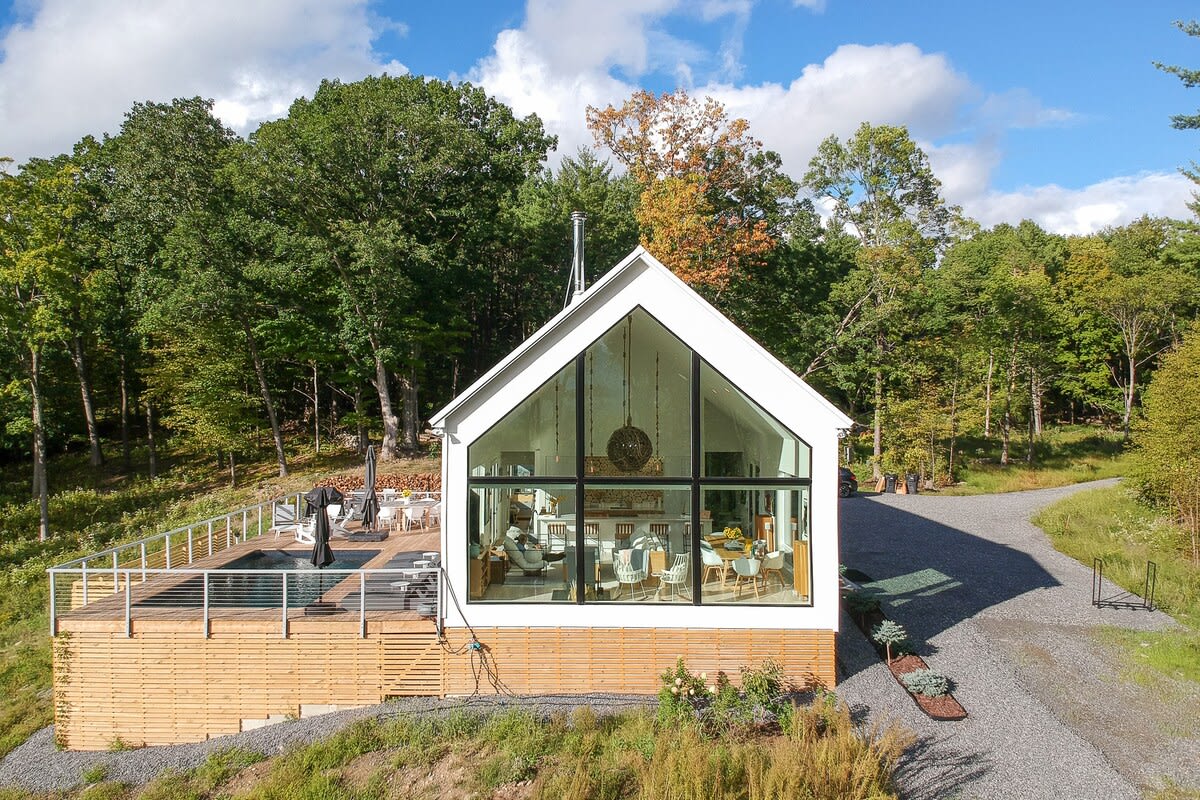 A striking modern A-frame house with a white exterior and large glass windows, situated on a hillside surrounded by lush green trees. A wooden deck with a swimming pool extends from the side of the house.