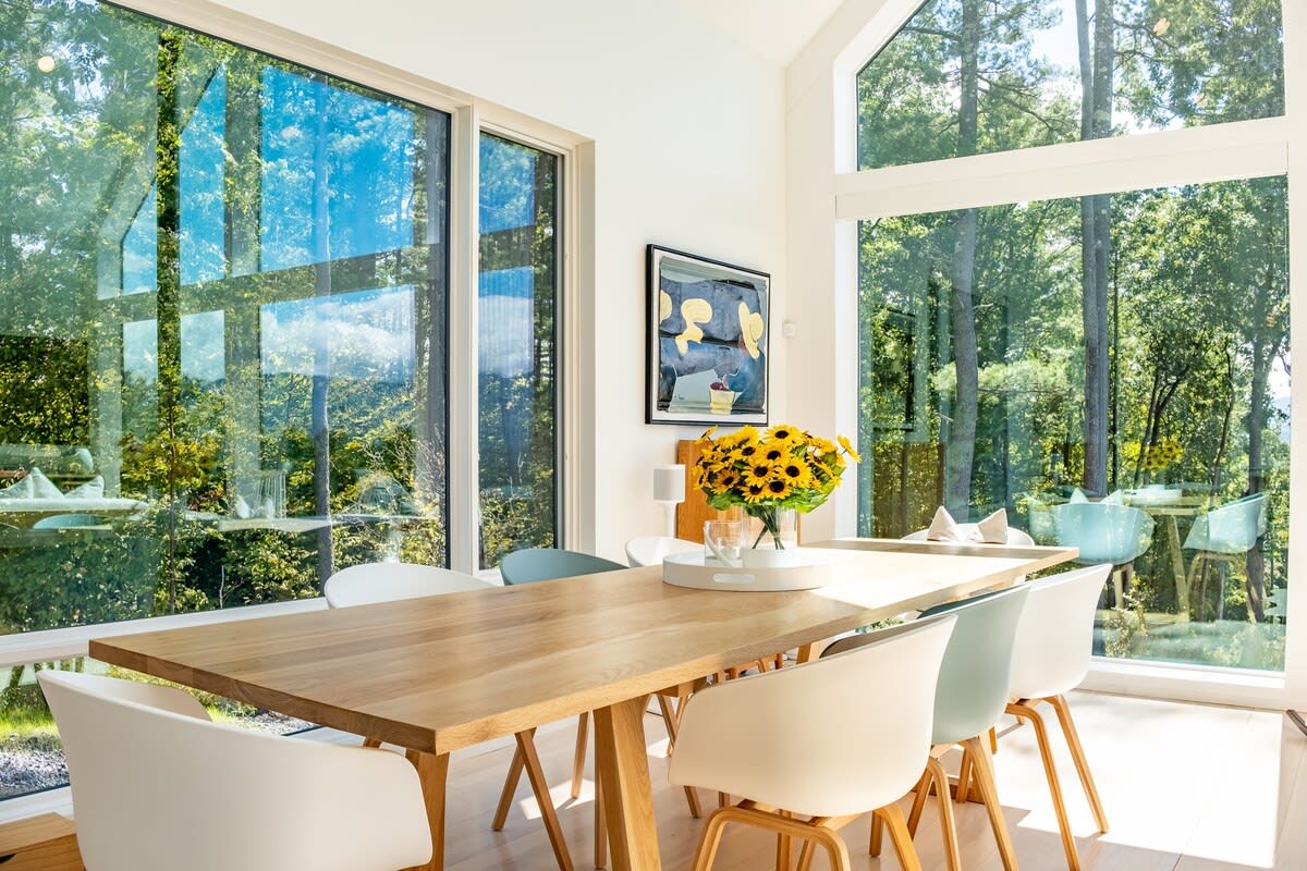 Bright dining area with a light wood table and modern chairs, surrounded by expansive floor-to-ceiling windows offering lush green forest views.