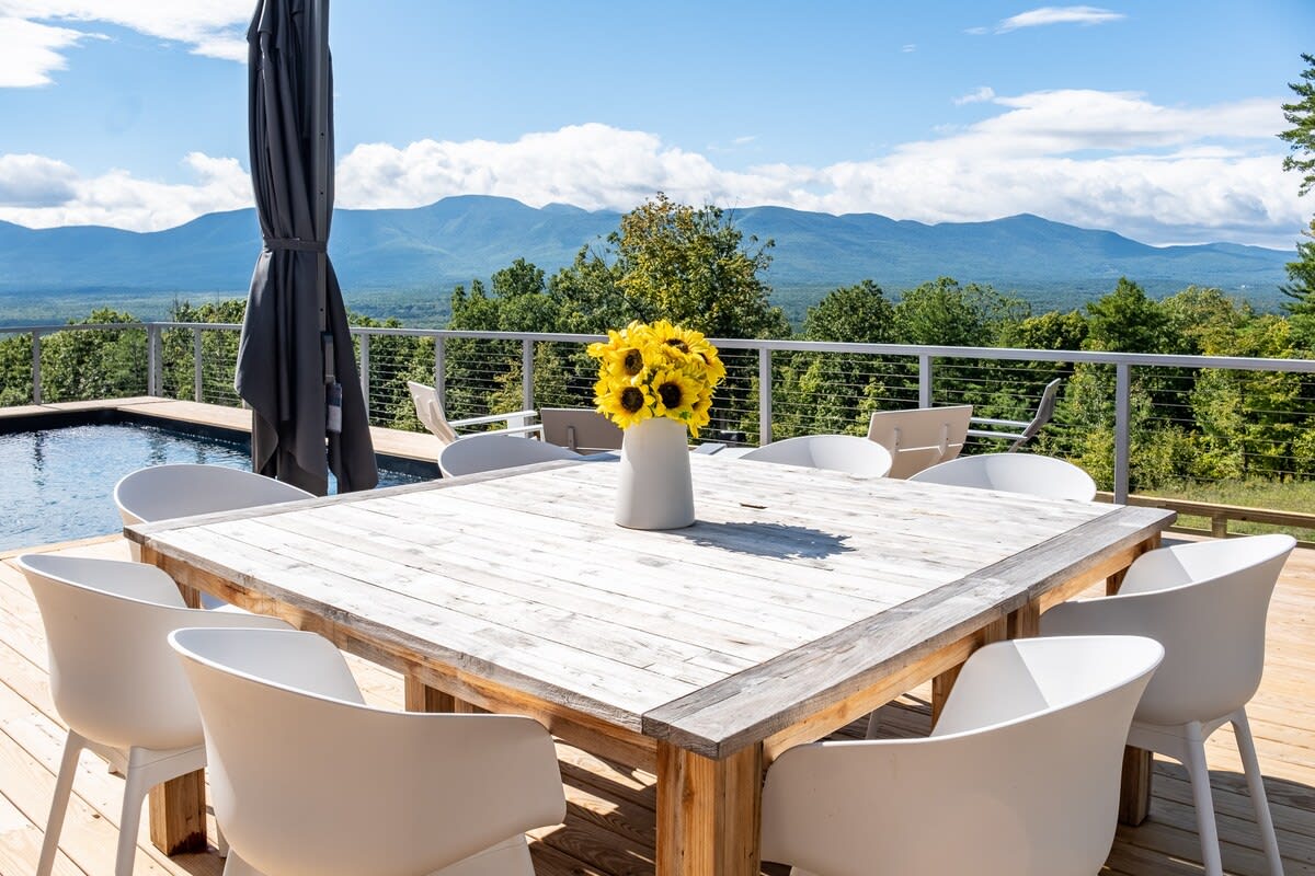 An outdoor dining area on a wooden deck, featuring a large rustic wooden table surrounded by white chairs, with a vase of sunflowers as a centerpiece. The scene is set against a stunning backdrop of distant mountains under a blue sky.