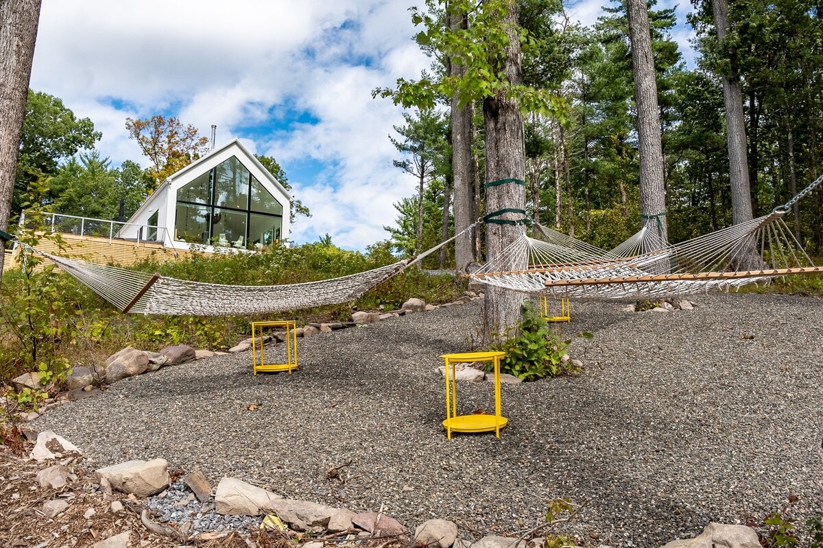 A serene outdoor relaxation area in a gravel yard, featuring multiple hammocks strung between tall trees and bright yellow side tables. The modern A-frame house with large windows is visible in the background.