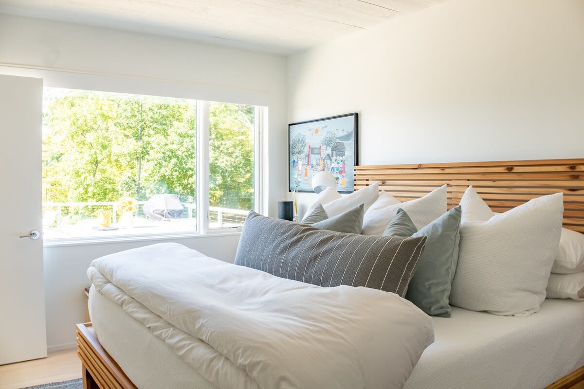 Serene bedroom with a wooden slatted headboard bed dressed in white and grey bedding, featuring a large window overlooking lush green trees and a glimpse of an outdoor balcony.