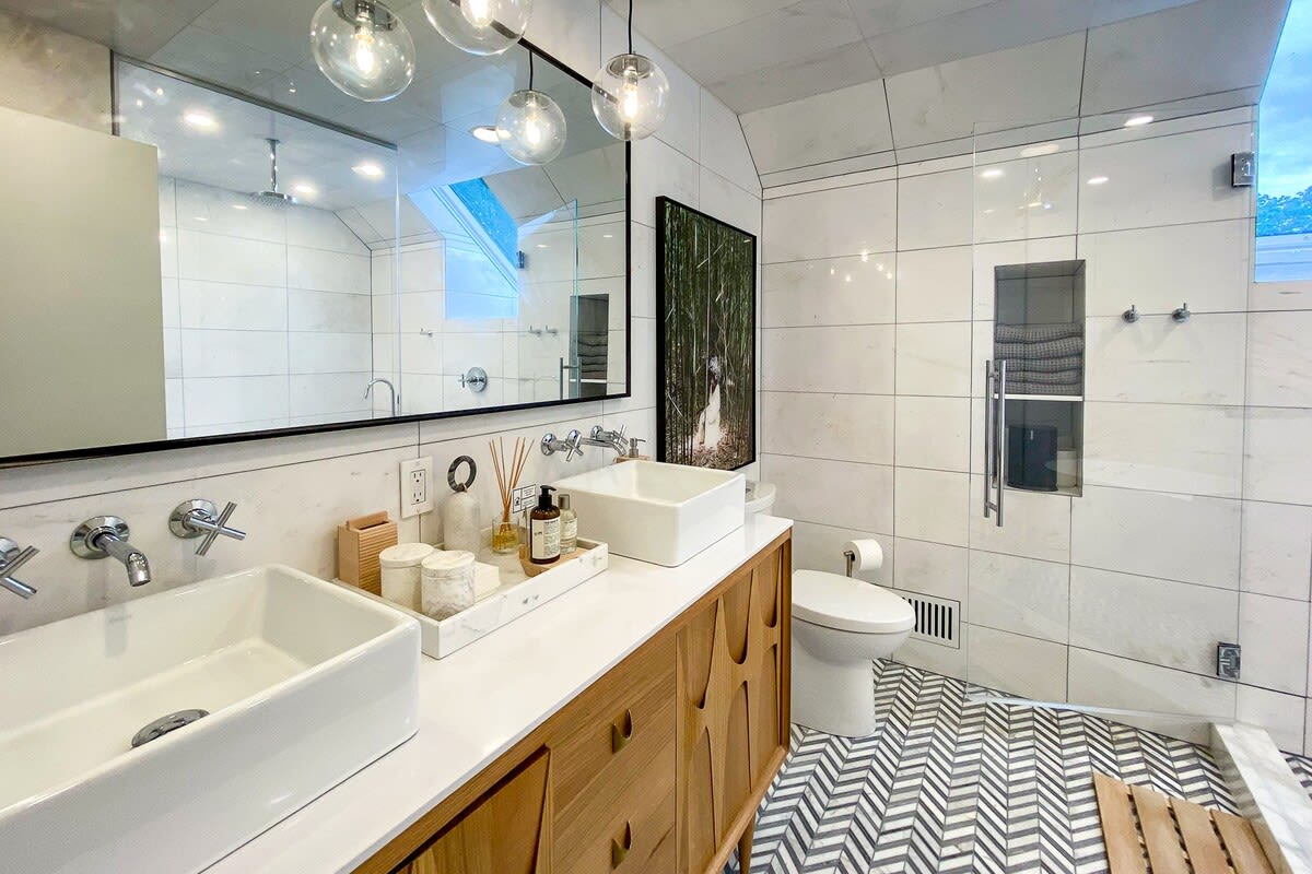 A stylish bathroom featuring a double vanity with white vessel sinks and a long mirror, set against white square wall tiles and a striking herringbone patterned floor. A glass-enclosed shower is visible on the right.
