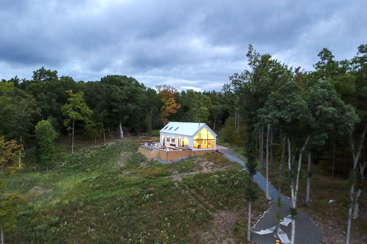 Aerial view of a modern white house with illuminated interiors and a large wooden deck, nestled in a clearing surrounded by dense forest under a dramatic cloudy sky. A gravel driveway leads to the house.