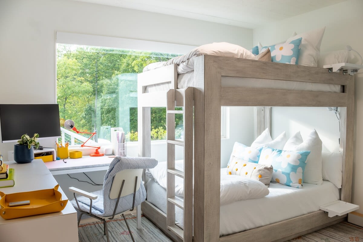 A bright bedroom featuring a light wood bunk bed with white bedding and colorful pillows, alongside a modern desk area with a computer monitor and a red task lamp, all set against a large window overlooking lush greenery.