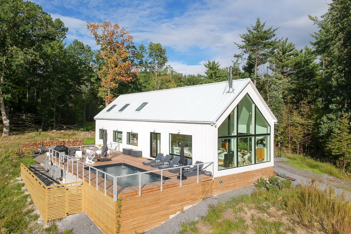 An aerial view showcasing the modern white A-frame house with a metal roof and skylights, nestled among green trees. A large wooden deck with a swimming pool is visible on the side, offering expansive views of the surrounding landscape.