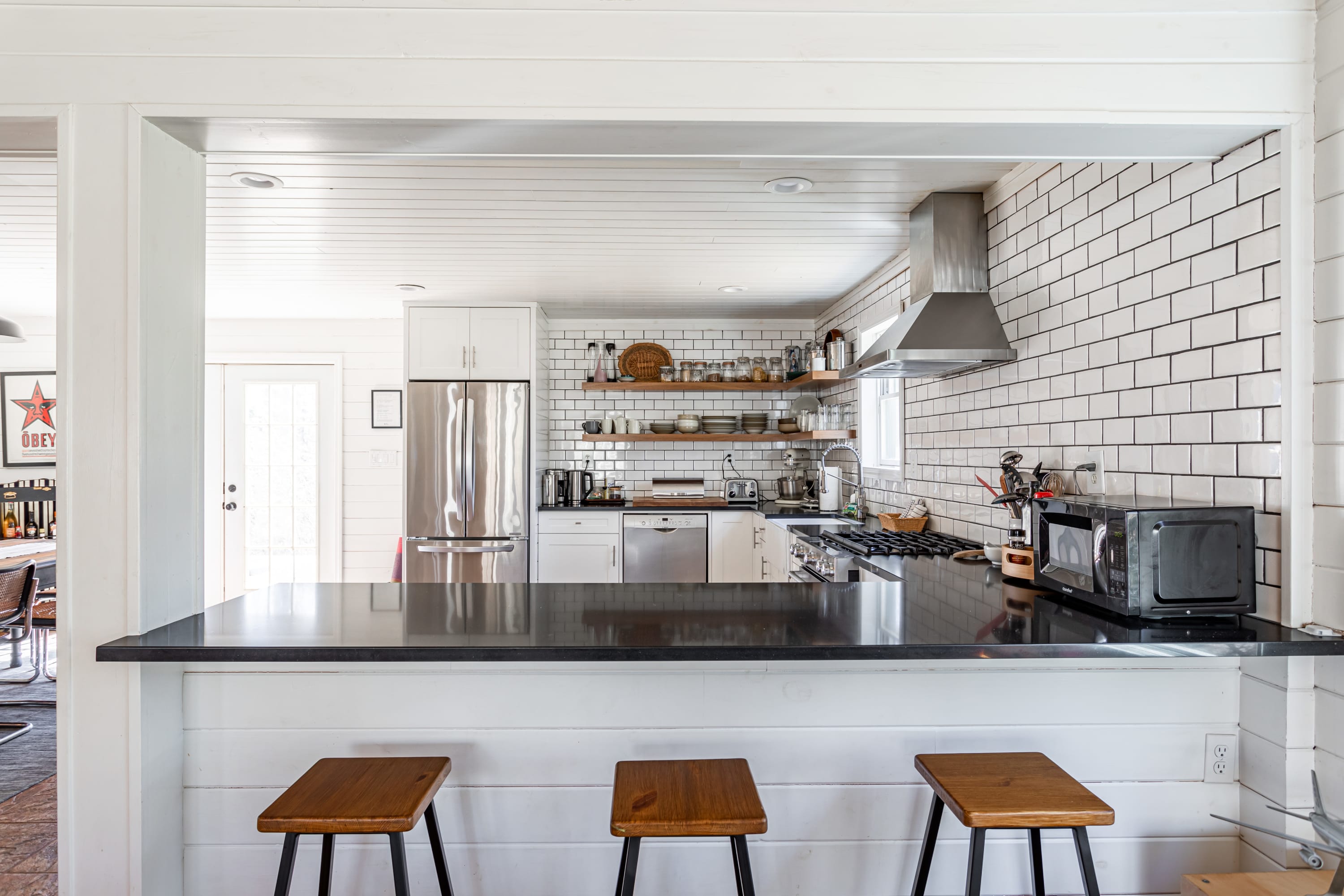 A view of the kitchen from a breakfast bar with three wooden stools, showcasing white subway tile backsplash, open shelving, and stainless steel appliances. The dark countertop provides a contrast to the white cabinetry.