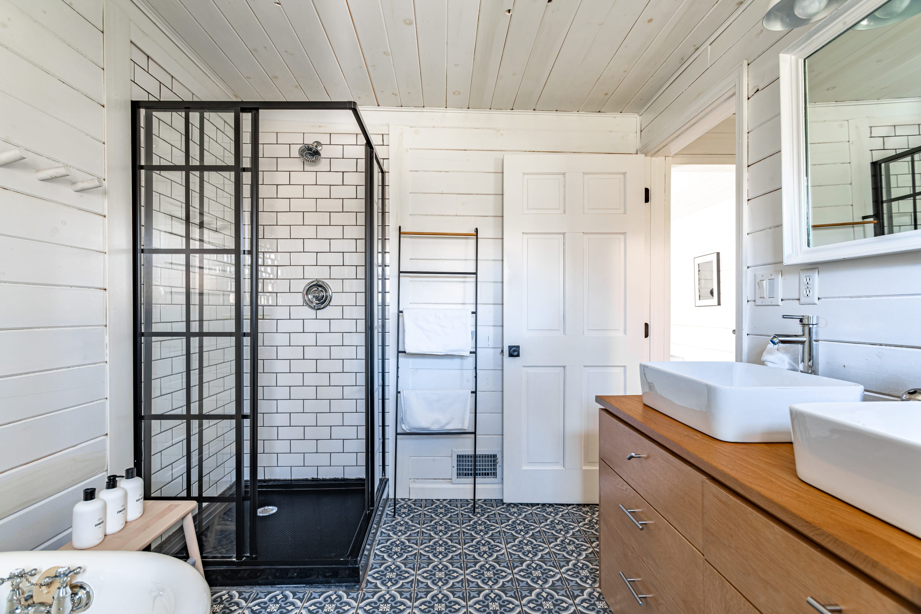 A modern bathroom with white shiplap walls and ceiling, featuring a black-framed glass shower enclosure with subway tiles, a freestanding clawfoot tub, and a wooden vanity with dual vessel sinks.