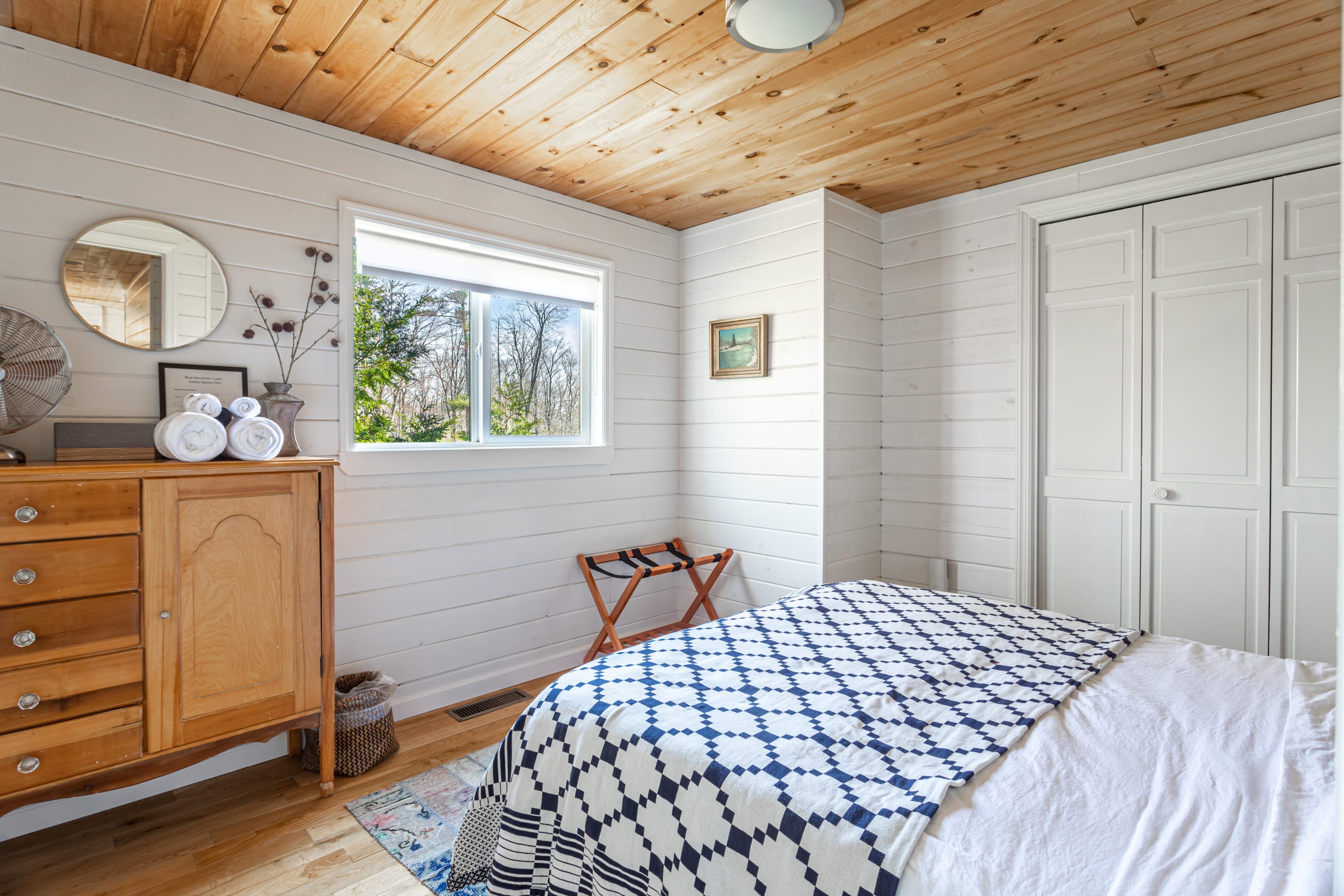Bright bedroom featuring white shiplap walls, a natural wood ceiling, and a bed with a blue and white quilt. A wooden dresser with a round mirror and a window overlooking trees are visible.