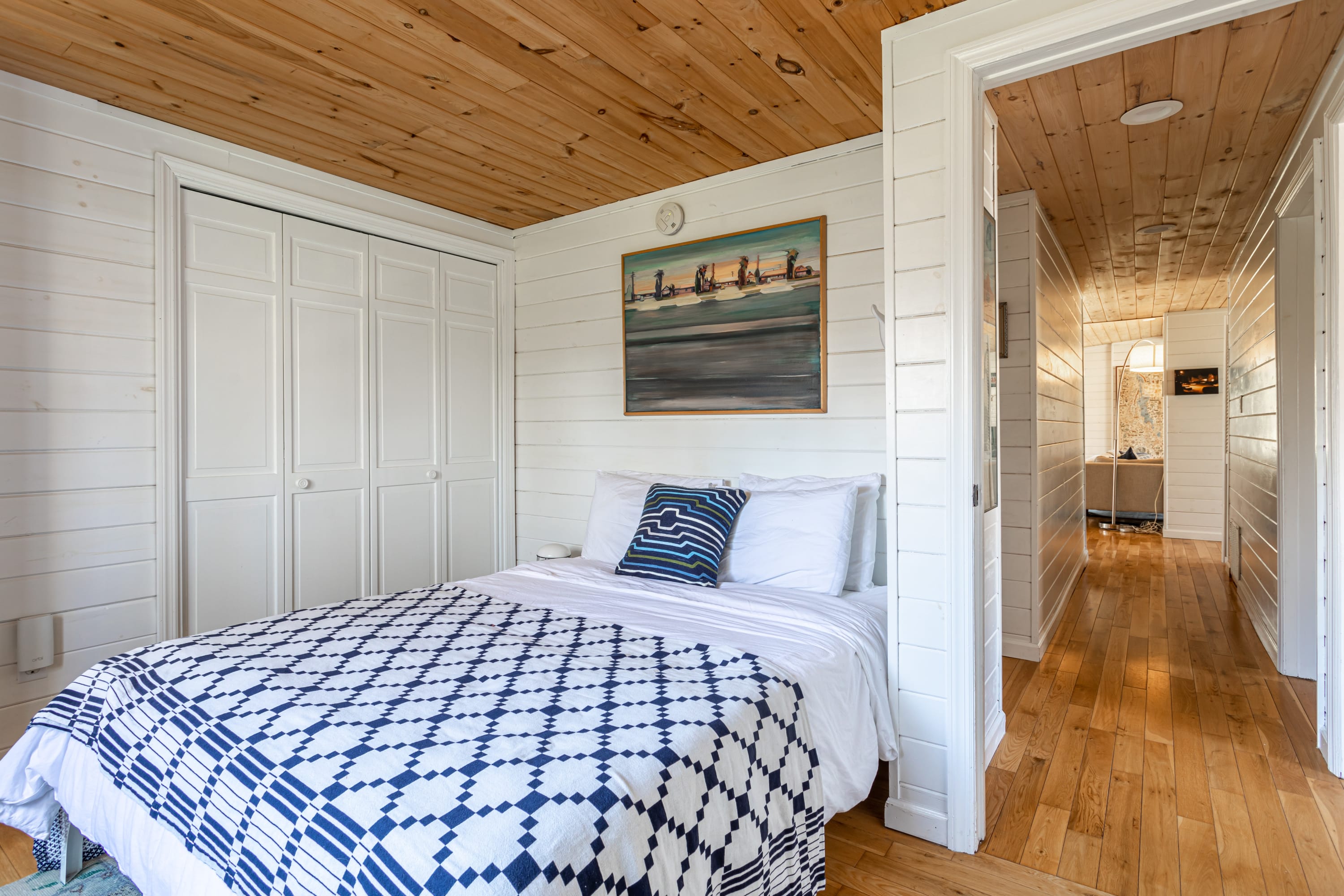 View of a bedroom with white shiplap walls, a wood plank ceiling, and a bed with blue and white patterned bedding. A doorway leads to a hallway with similar wood and shiplap finishes.