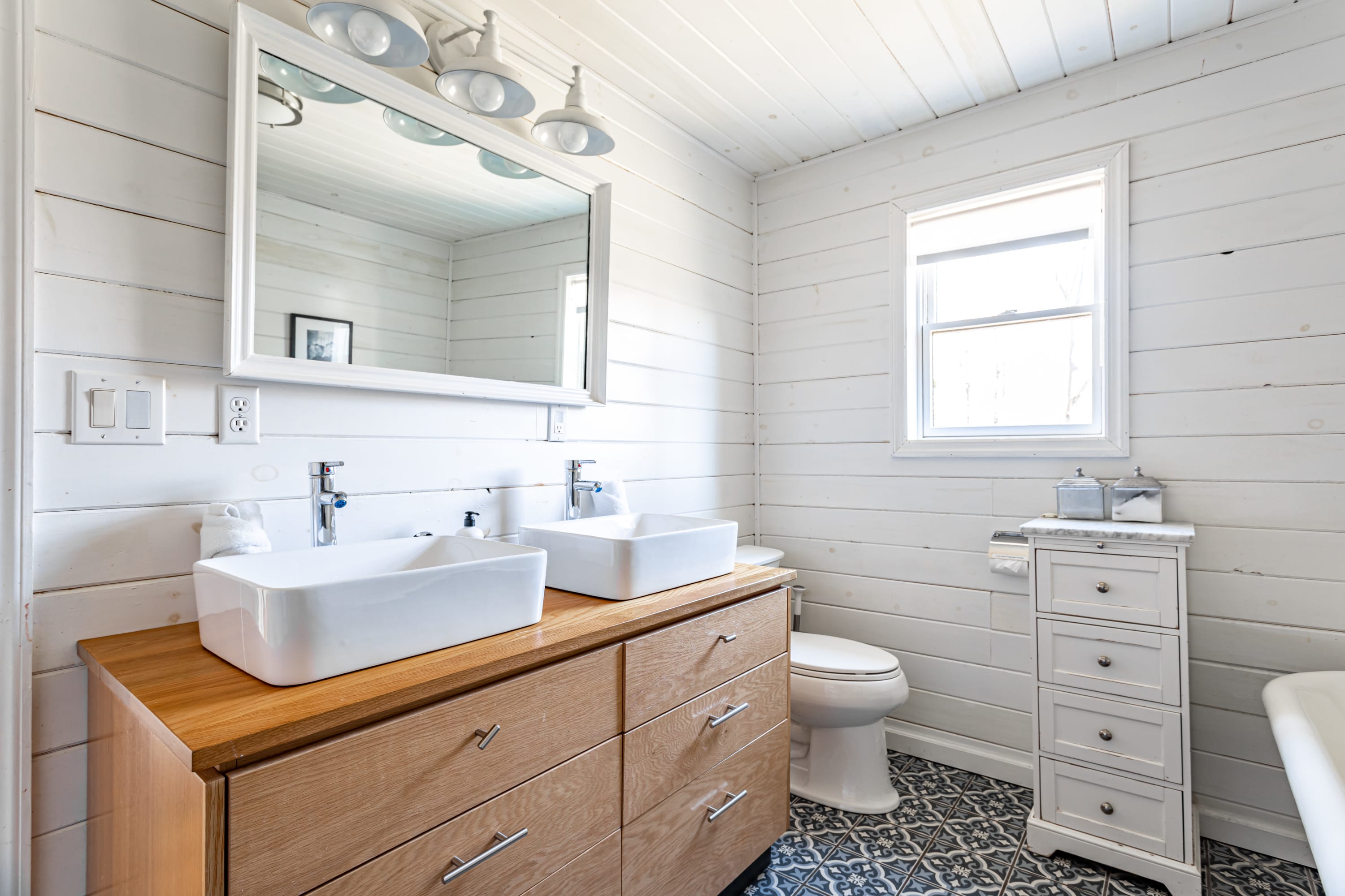 The bathroom's wooden vanity with dual white vessel sinks and a large mirror, a toilet, and a small white storage cabinet, all set against white shiplap walls and distinctive patterned floor tiles.