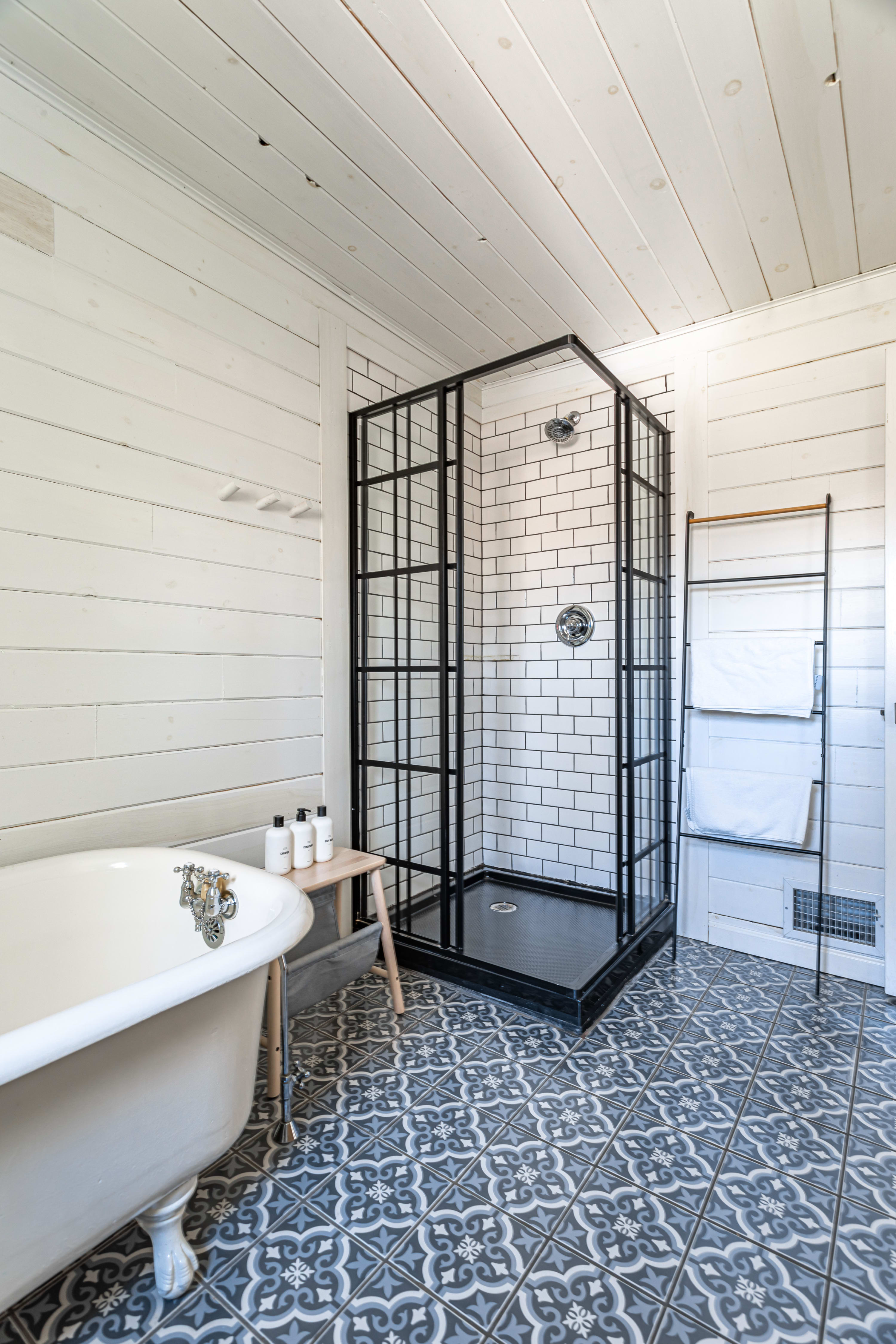 A detailed view of the bathroom's black-framed glass shower enclosure with white subway tiles and a black base, alongside a classic white clawfoot tub, set against white shiplap walls and patterned floor tiles.