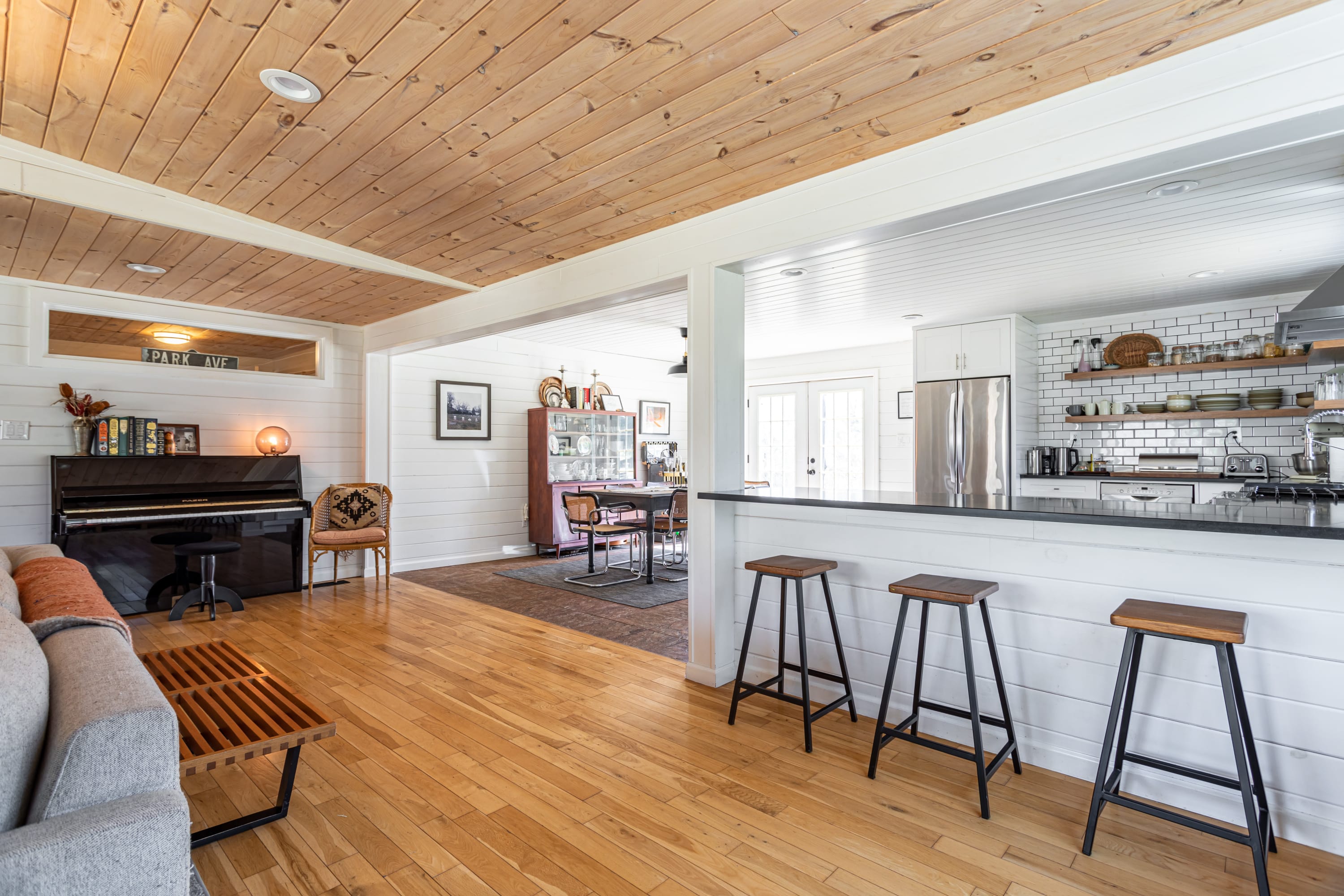 An open-concept living space with light wood floors and a wood-paneled ceiling, transitioning into a kitchen with a breakfast bar and bar stools, featuring white shiplap walls and a piano in the foreground.