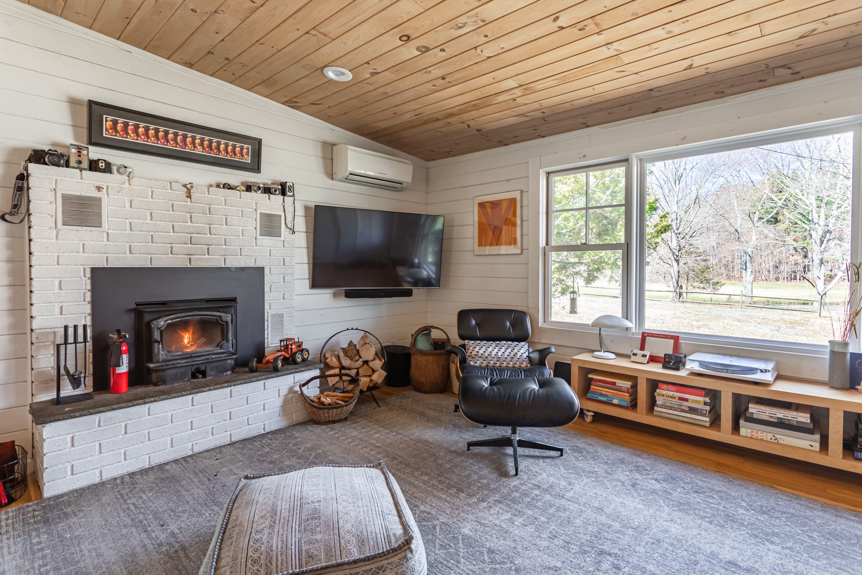 A corner of the living room featuring a white brick fireplace with a wood-burning insert, a flat-screen TV mounted above, and an Eames lounge chair. A large window offers views of the bare trees outside.