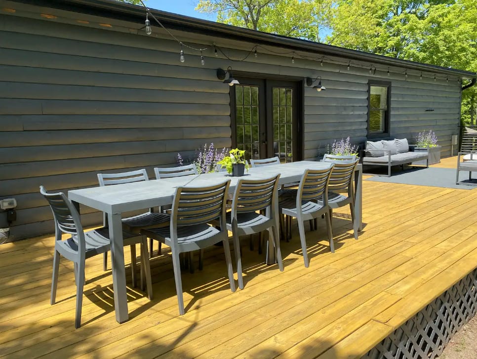 A long gray outdoor dining table with matching chairs is set on a golden wooden deck, adjacent to the dark-paneled house with string lights. Planters with purple flowers add a touch of color.