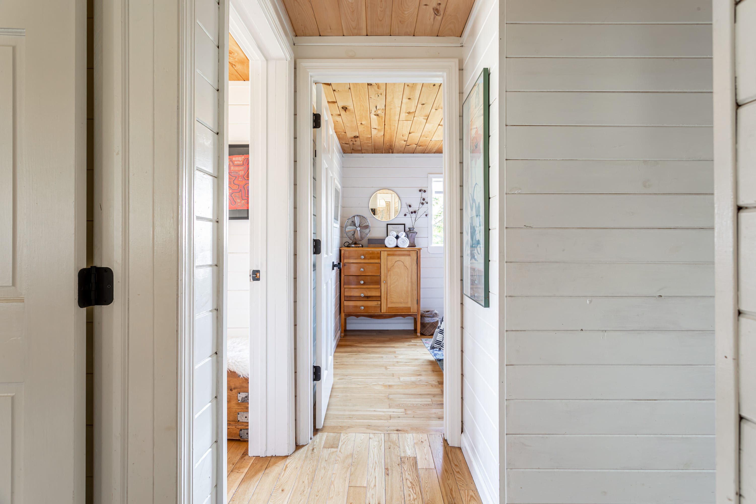 A bright hallway with white shiplap walls and light wood floors and ceiling, leading into a bedroom with a wooden dresser and round mirror. White doors line both sides of the hall.