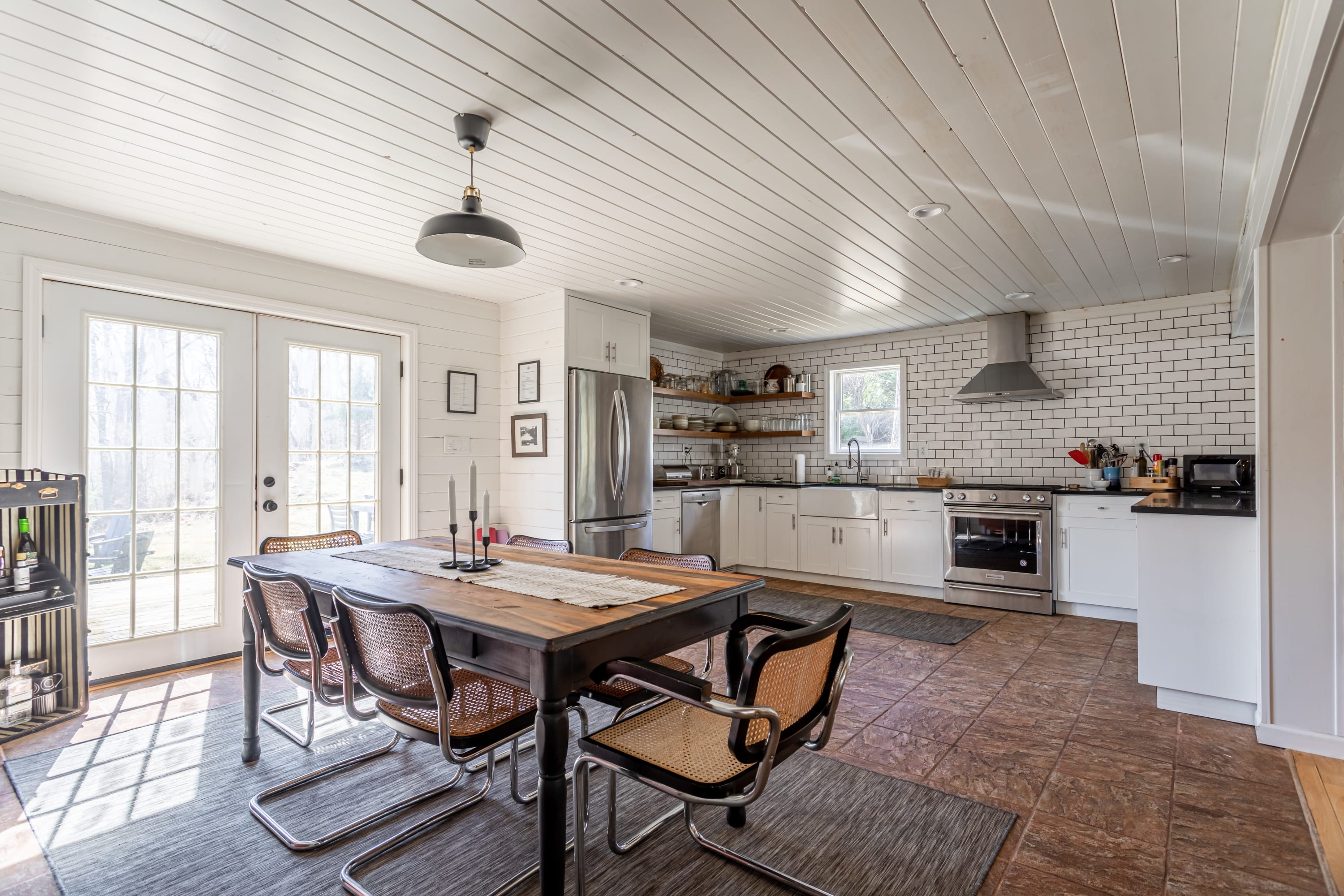 An open-concept kitchen and dining area featuring white shaker cabinets, a farmhouse sink, stainless steel appliances, and white subway tile backsplash. The dining table with cane chairs is on a tiled floor, adjacent to French doors.