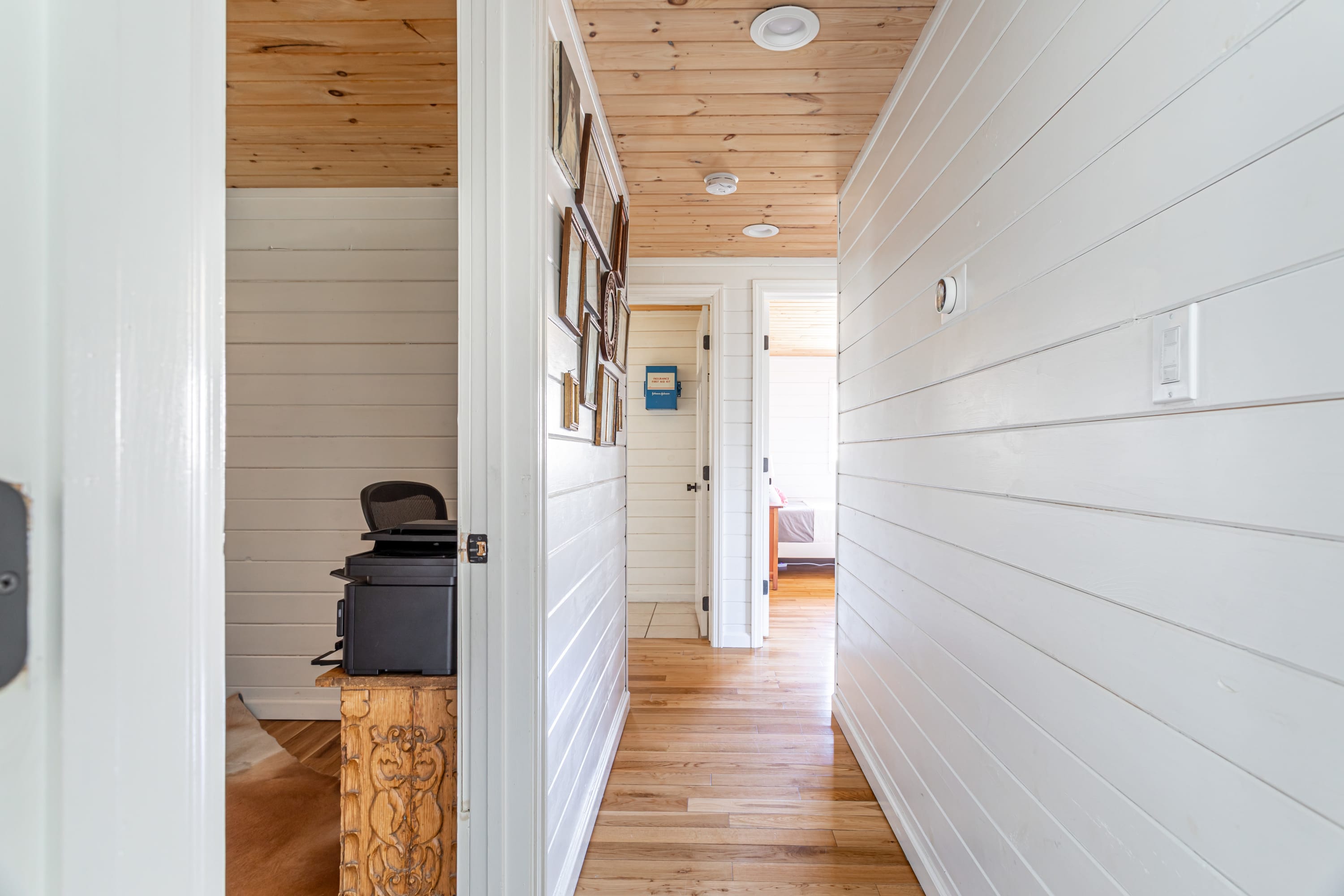 A bright hallway with white shiplap walls and light wood flooring, leading to several rooms, with a wooden ceiling and framed pictures adorning one wall.