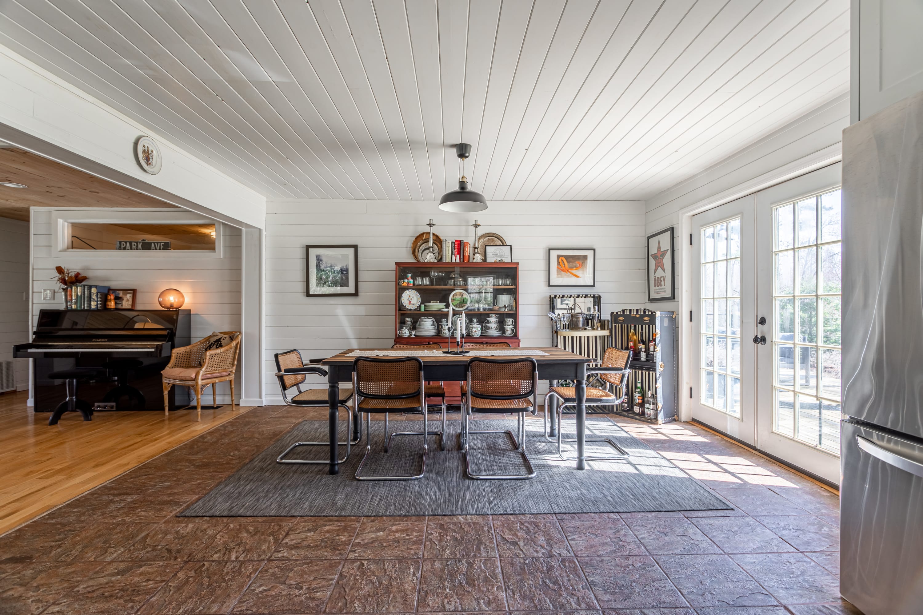 A spacious dining area with a dark wood table and six cane-back chairs, set on a tiled floor. French doors lead to the outside, and a white shiplap ceiling with a pendant light completes the rustic modern look.