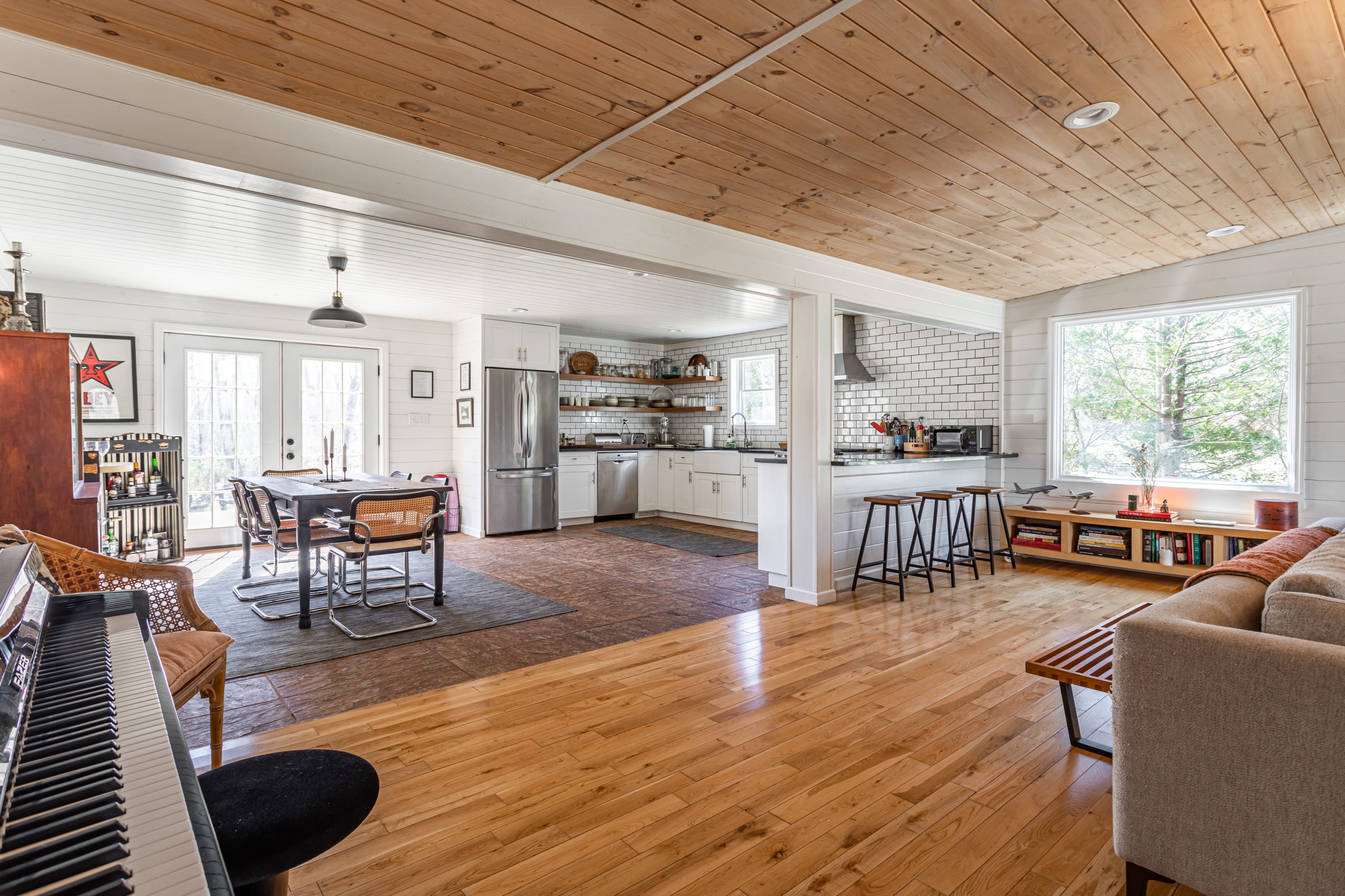An open-concept living space with hardwood floors, a dining area with a table and chairs, and a view into the kitchen. The room features white shiplap walls and a natural wood plank ceiling, with a piano in the foreground.