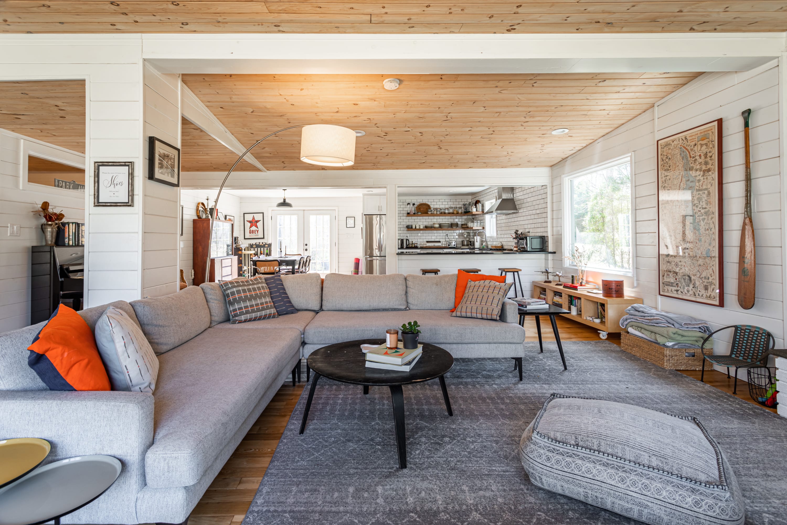 An open-concept living space with a large L-shaped sectional sofa, a coffee table, and a view into the adjacent kitchen area with a breakfast bar. The room features white shiplap walls and a light wood plank ceiling.