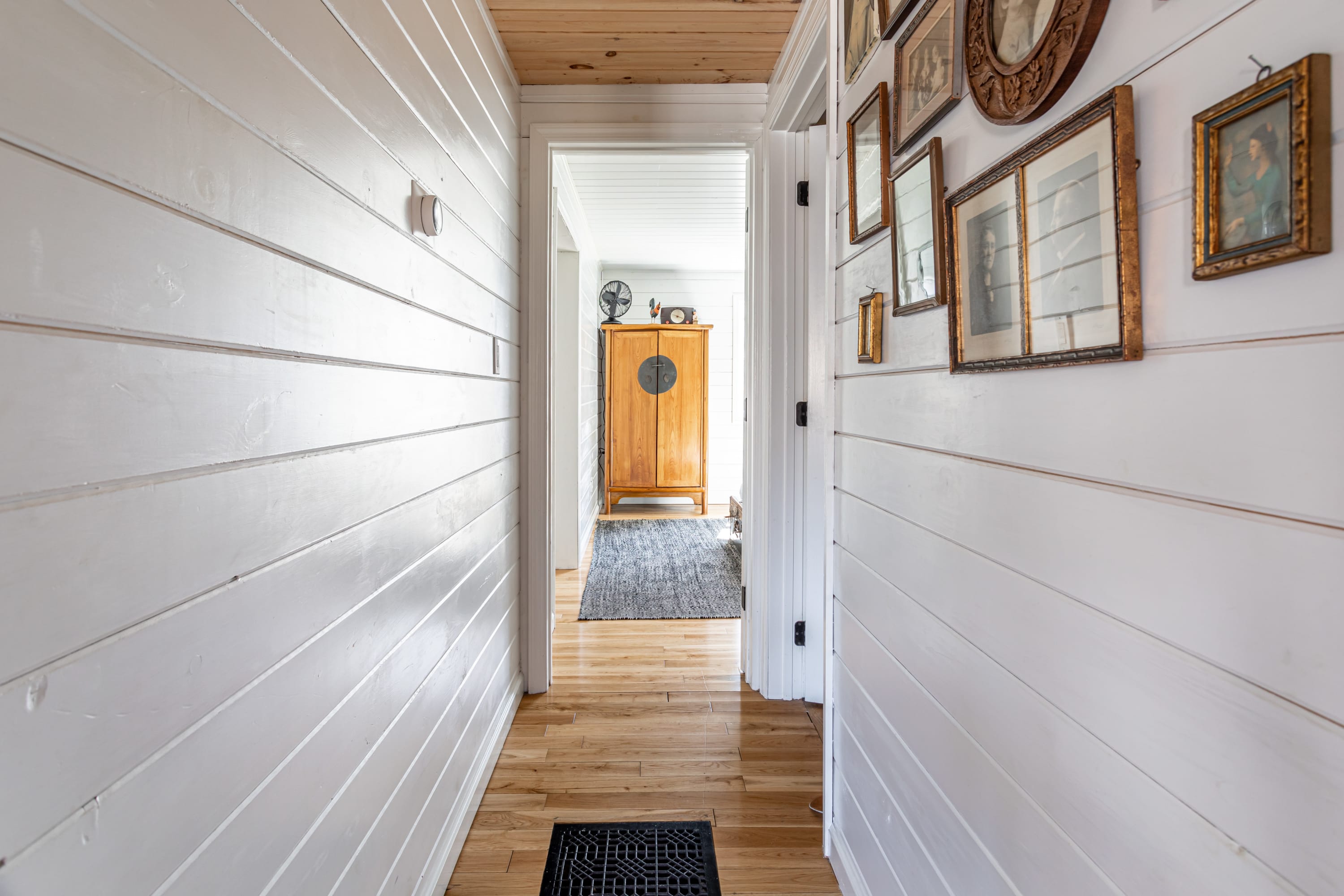 A narrow hallway with white shiplap walls and light wood flooring, adorned with framed artwork, leading to an open doorway revealing another room with a wooden cabinet.