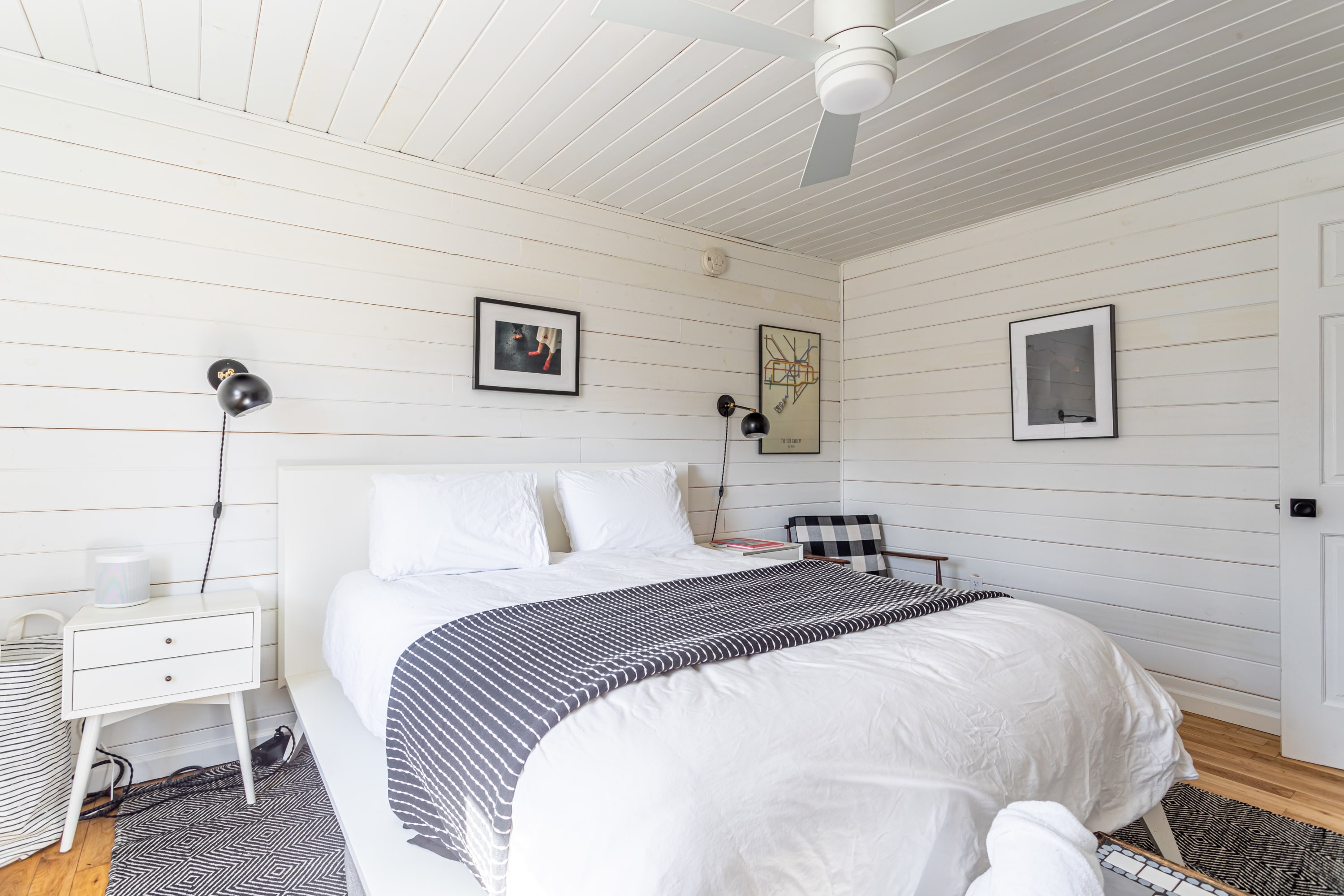 A cozy bedroom with white shiplap walls and ceiling, featuring a white platform bed with white and striped bedding, a white nightstand, and a modern ceiling fan, with framed art on the walls.