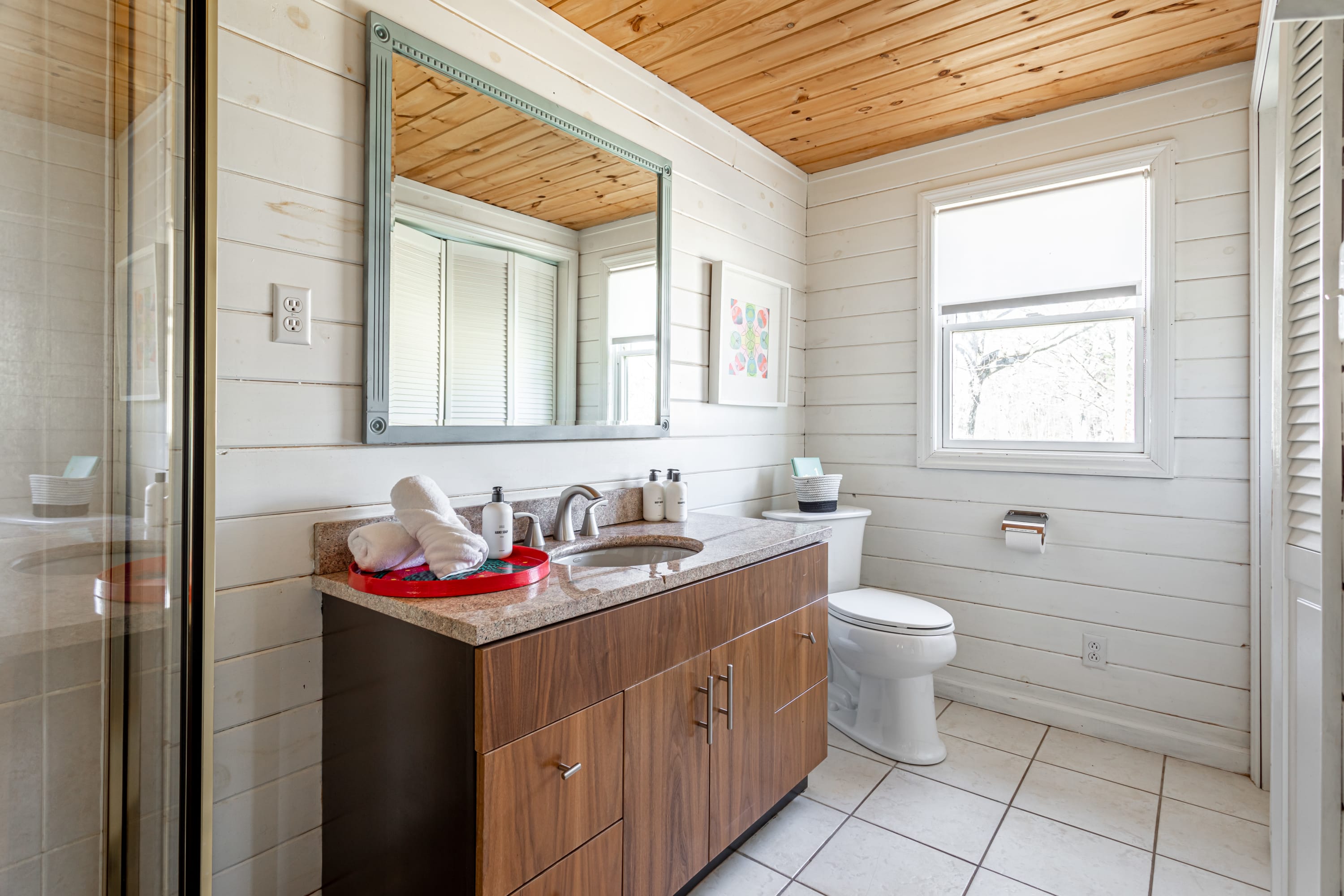 Bright bathroom with white shiplap walls, a wood ceiling, and a window. It features a modern vanity with a granite top, a toilet, and a large mirror with a light blue frame.