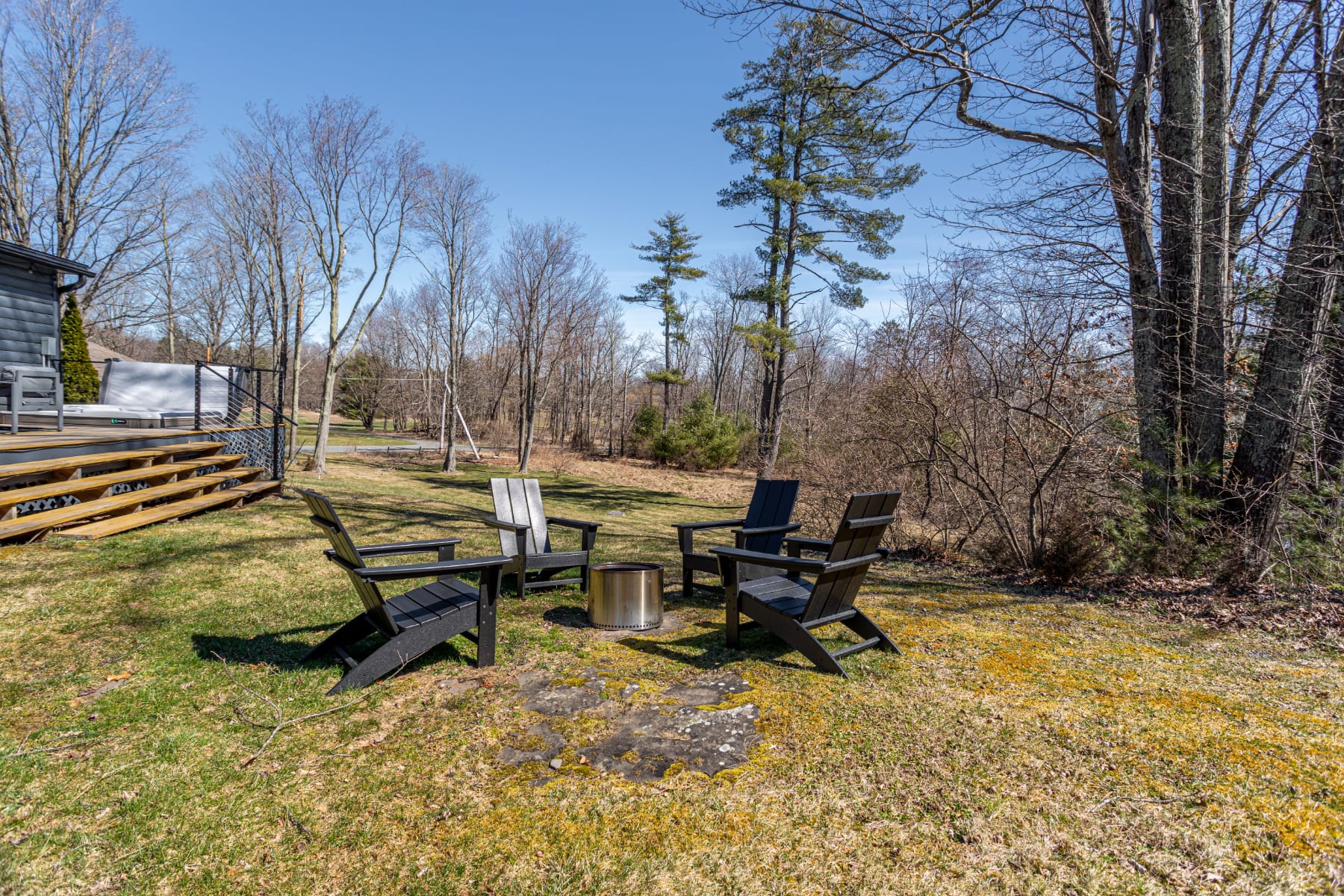 A circular fire pit area with four black Adirondack chairs is set on a grassy lawn, surrounded by bare trees in a natural, wooded setting. The deck with the hot tub is visible in the background.