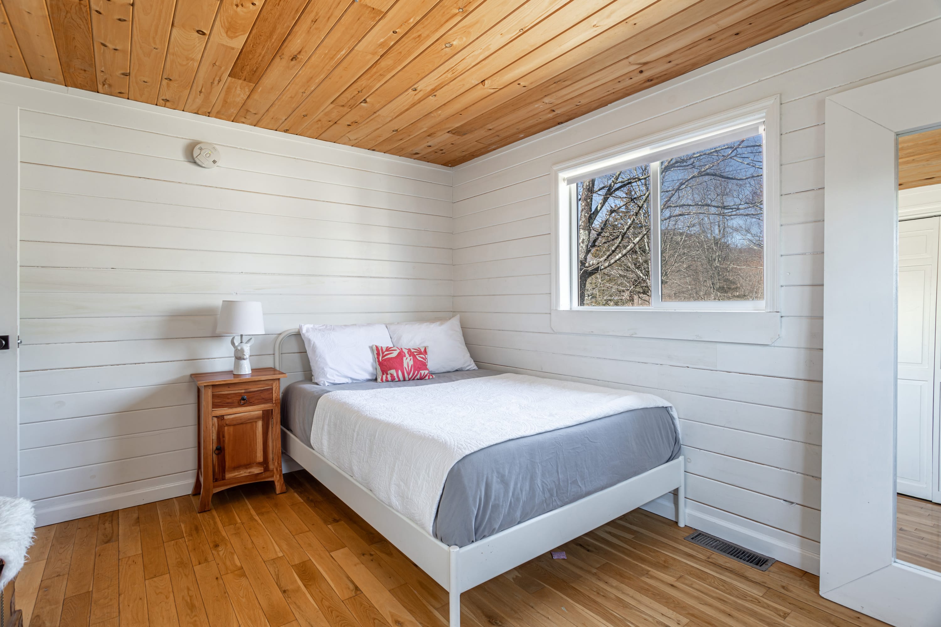 Cozy bedroom with white shiplap walls, a wood plank ceiling, and a white bed with a grey and white quilt. A small wooden nightstand with a lamp and a window are visible.