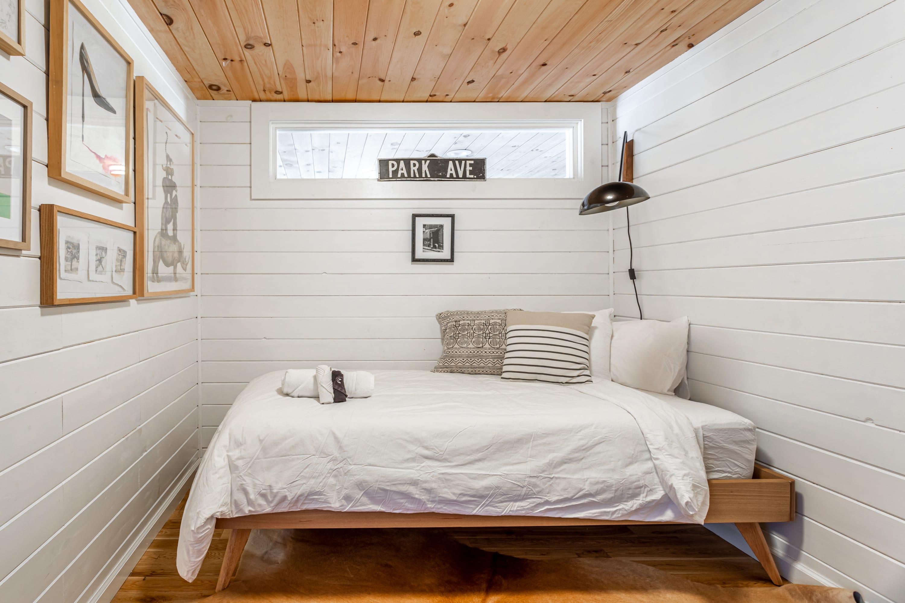 Small bedroom featuring white shiplap walls, a wood plank ceiling, and a single bed with white bedding. A narrow window and a "PARK AVE" sign are above the bed, with framed artwork on the adjacent wall.