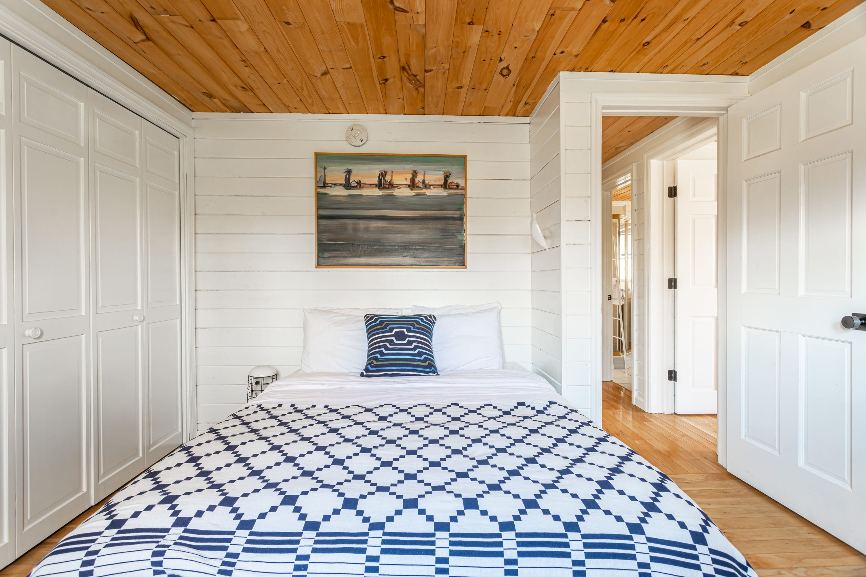 Cozy bedroom with white shiplap walls, a light wood plank ceiling, and a bed with a blue and white geometric patterned quilt. A painting hangs above the headboard, and white bi-fold closet doors are visible.