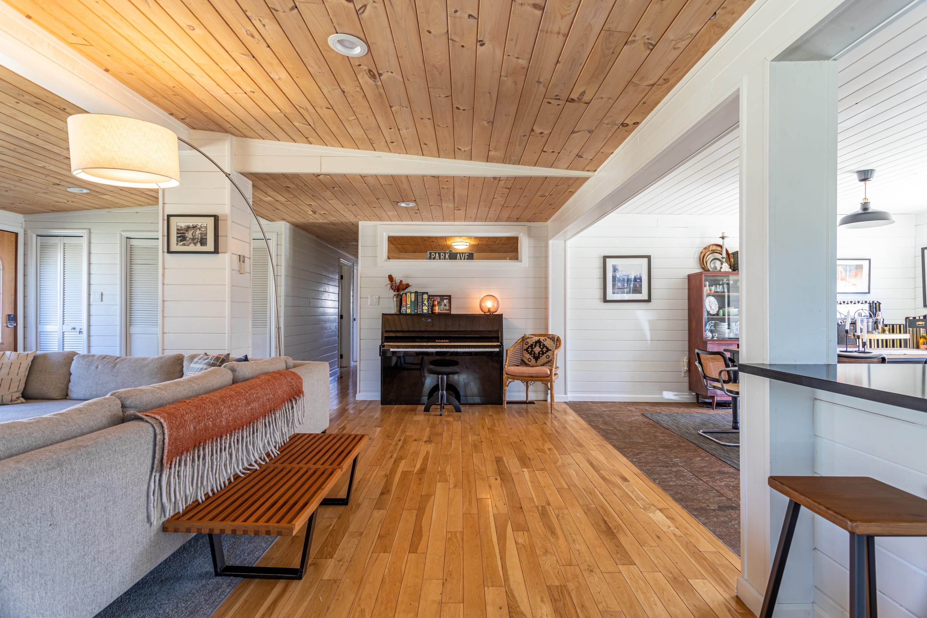 A wide view of the open-concept space, showing the living room with a sectional sofa transitioning to a dining area and kitchen. Light hardwood floors extend through the living space, with a piano visible in the background.