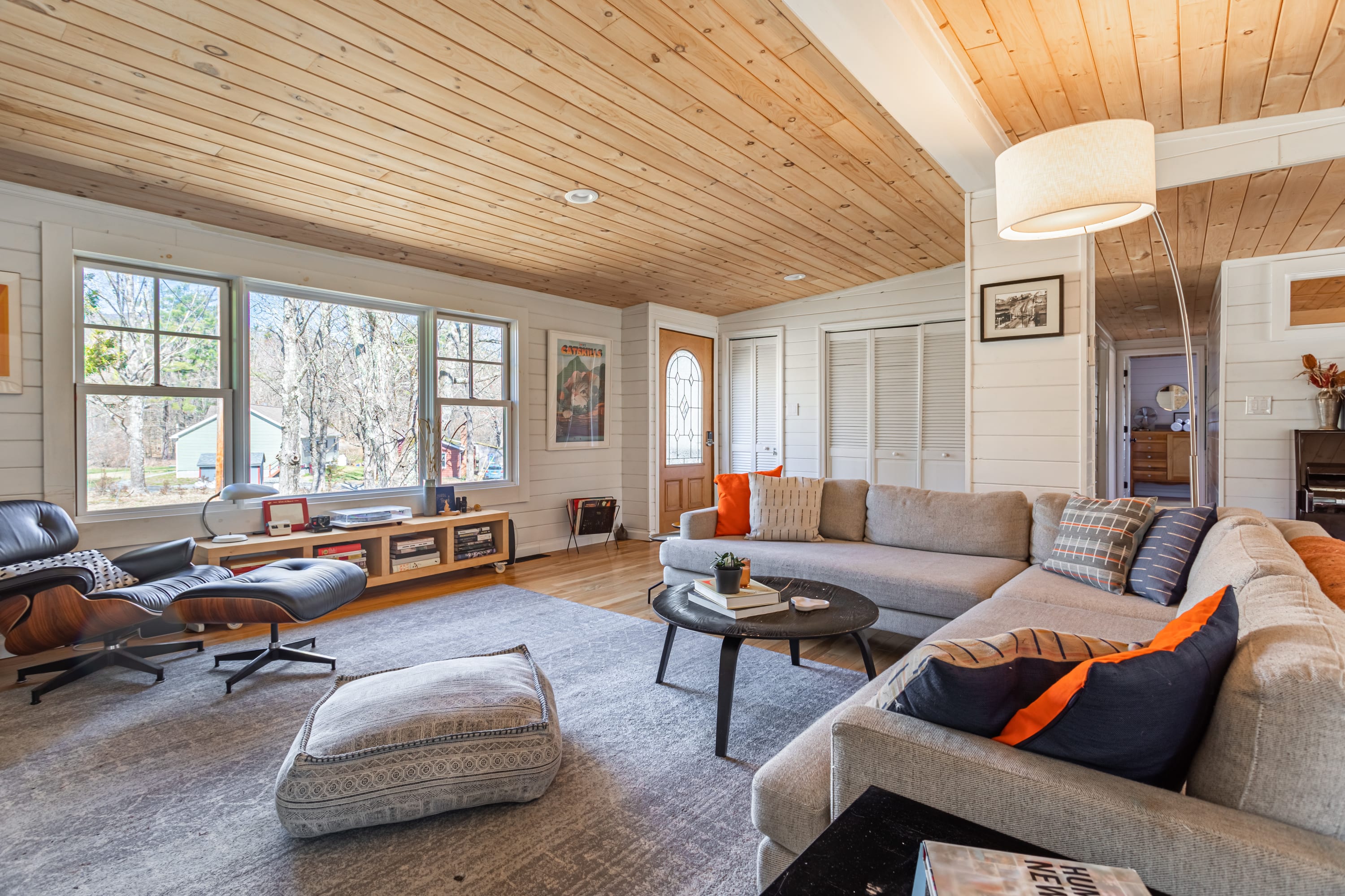 A bright living room with a large L-shaped sectional sofa and an Eames lounge chair, facing a large window overlooking the outdoors. The room has white shiplap walls, a light wood plank ceiling, and light hardwood floors.