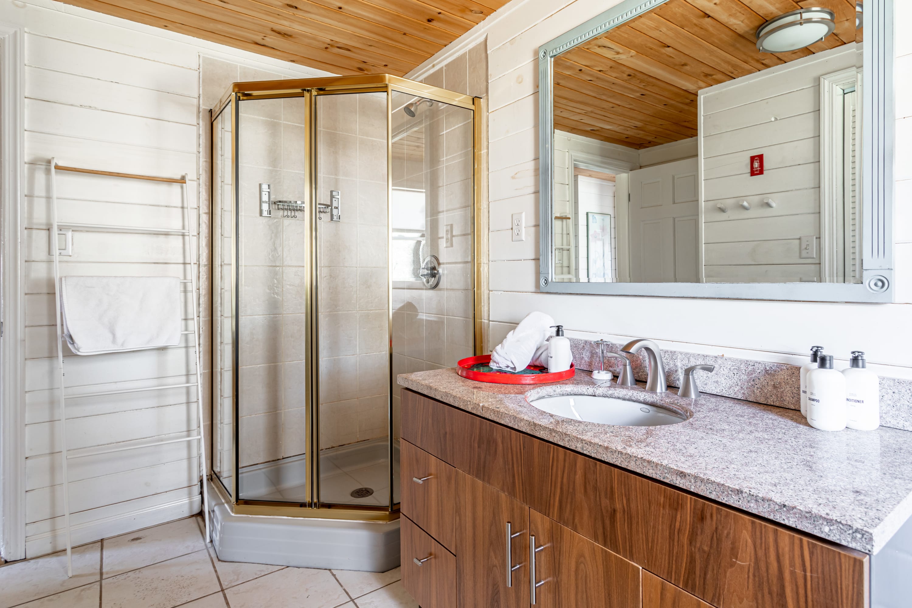 Bathroom with white shiplap walls, a wood plank ceiling, and a corner glass shower. A modern vanity with a granite countertop and a large mirror are also visible.