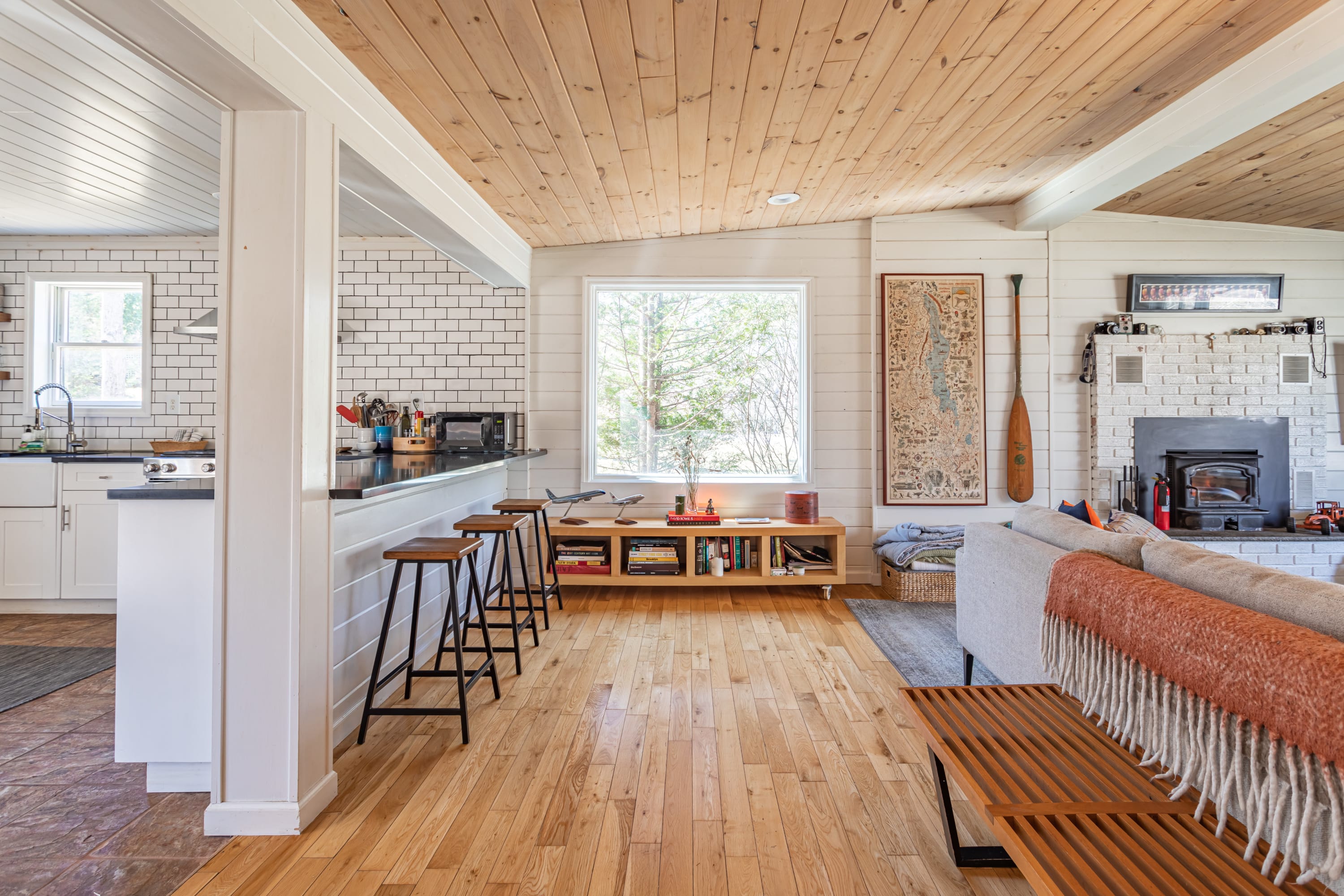 A view from the living room towards the kitchen, showing a breakfast bar with three stools and light hardwood floors. The kitchen features white subway tile backsplash and a sink under a window.
