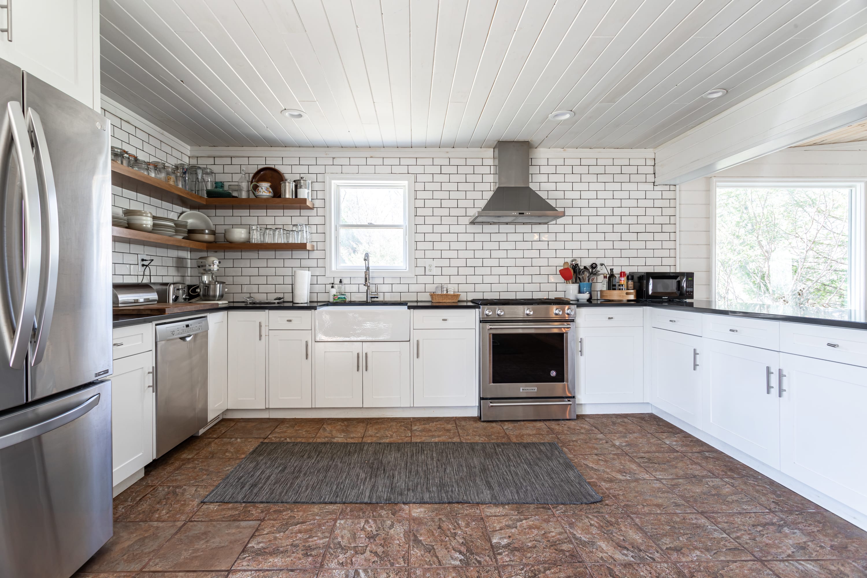 A bright, modern farmhouse-style kitchen featuring white subway tile backsplash, open wooden shelving, white shaker cabinets, dark countertops, and stainless steel appliances, including a large farmhouse sink.