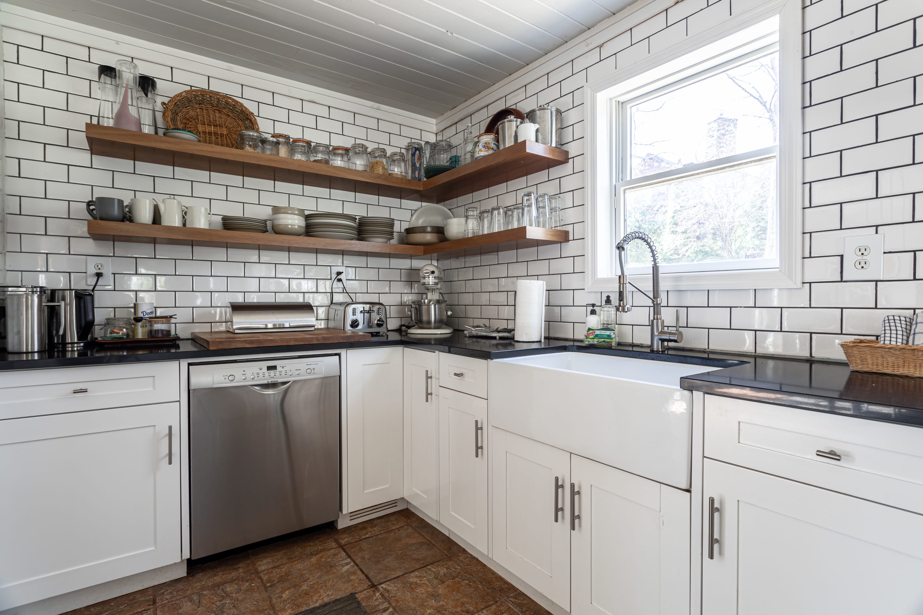A detailed view of the modern farmhouse kitchen, showcasing white subway tile backsplash, open wooden shelves filled with dishes, a stainless steel dishwasher, and a large white farmhouse sink with a pull-down faucet.