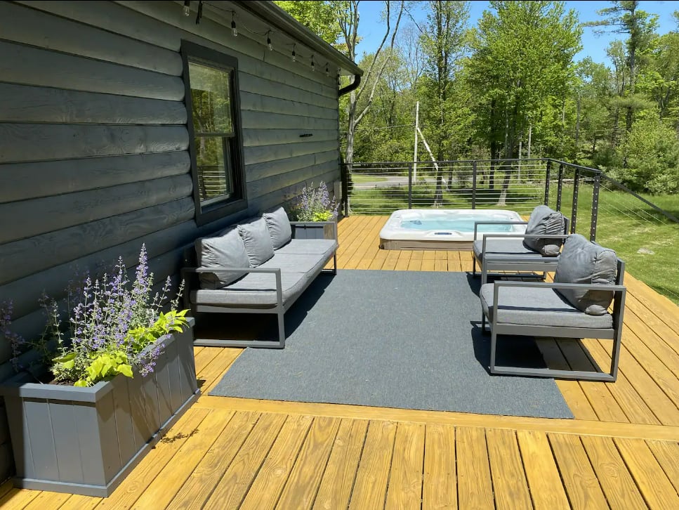 A modern gray outdoor lounge set, including a sofa and two chairs, is arranged on a dark gray rug atop a golden wooden deck. Planters with purple flowers line the house, with a hot tub visible in the background.