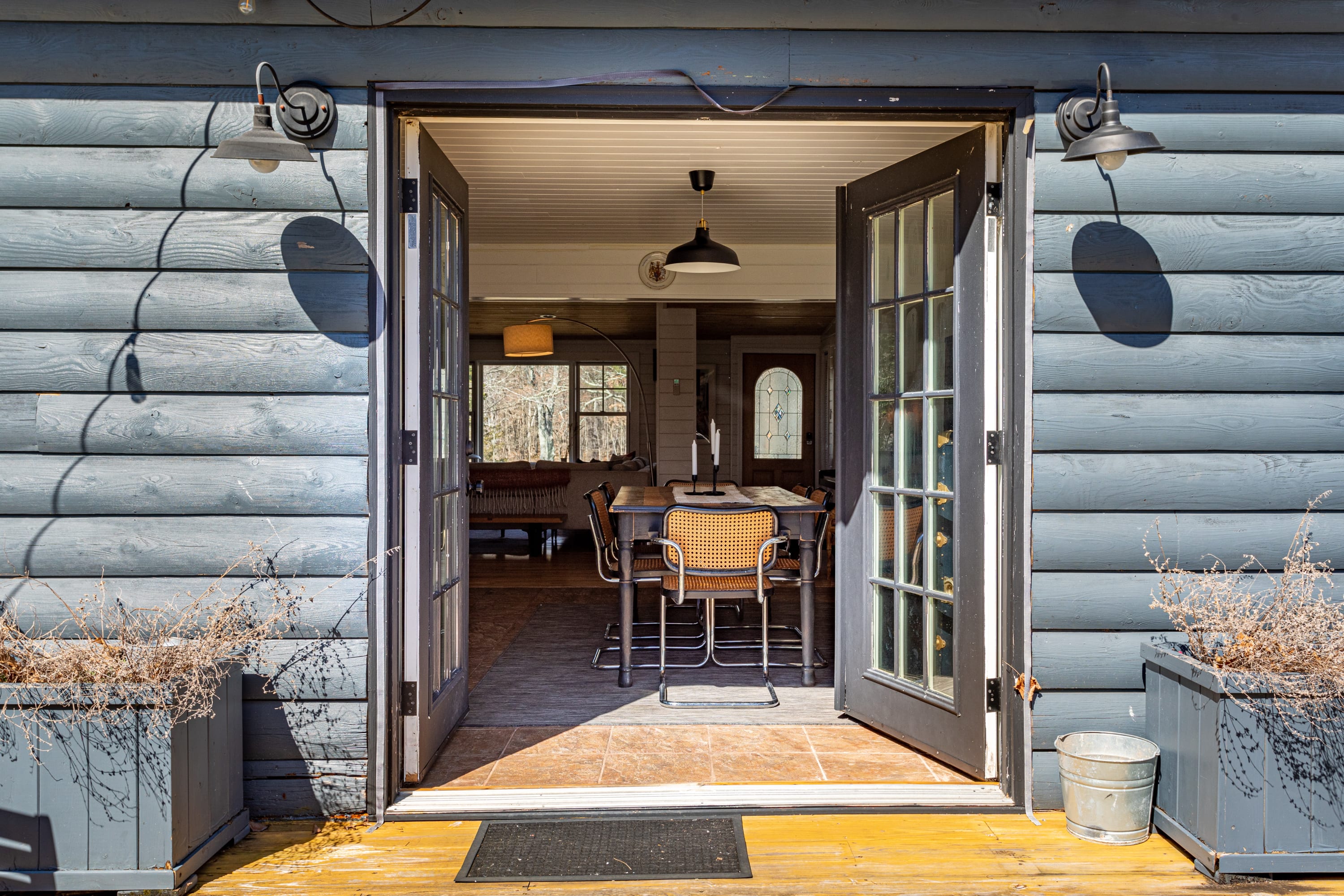 Double French doors open to reveal an indoor dining area with a dark table and chairs, set against white shiplap walls and a wood plank ceiling. The exterior features dark gray siding and outdoor sconces.